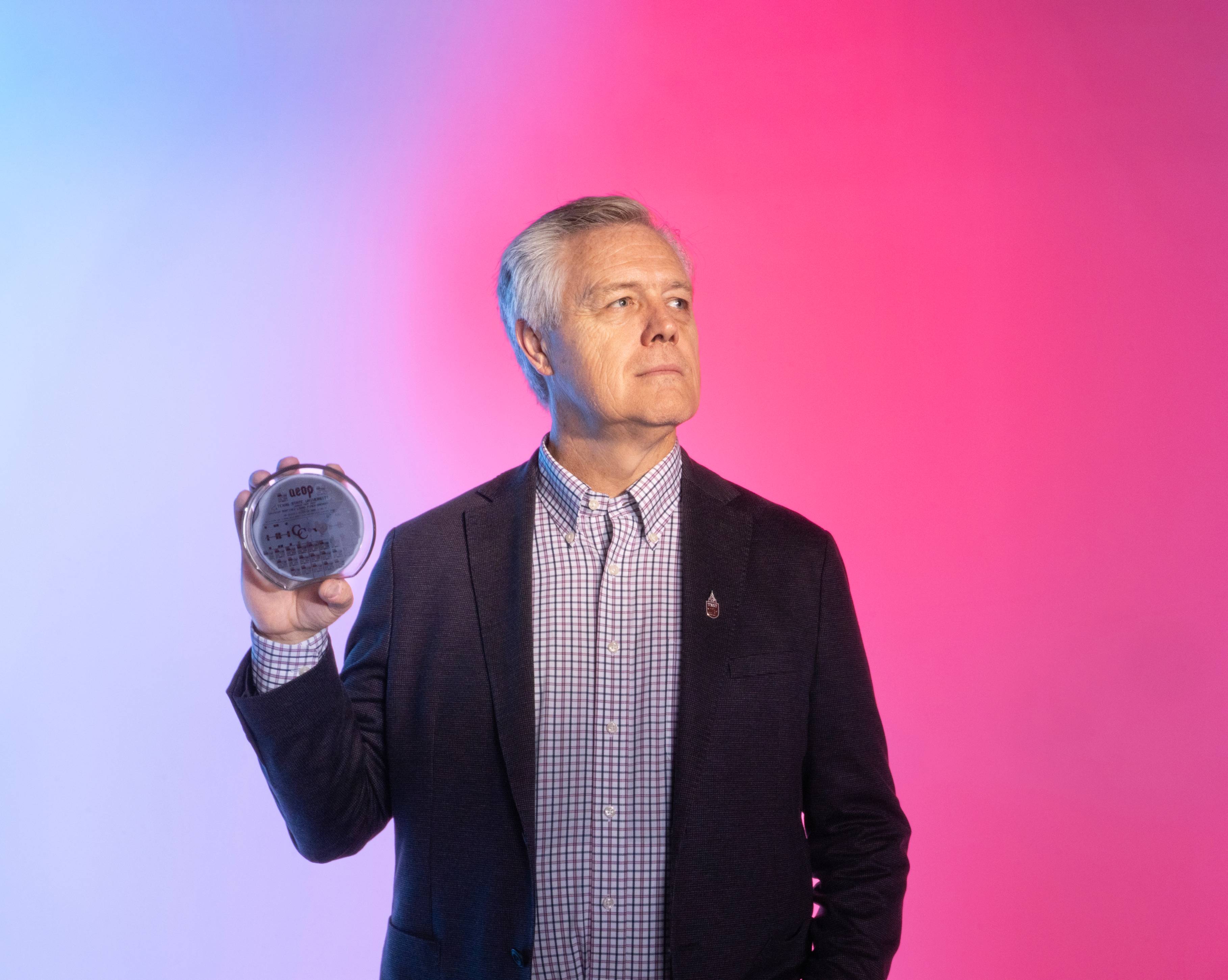 Dr. Edwin Piner holds up a semiconductor in his right hand while standing in front of a pink and blue gradient background