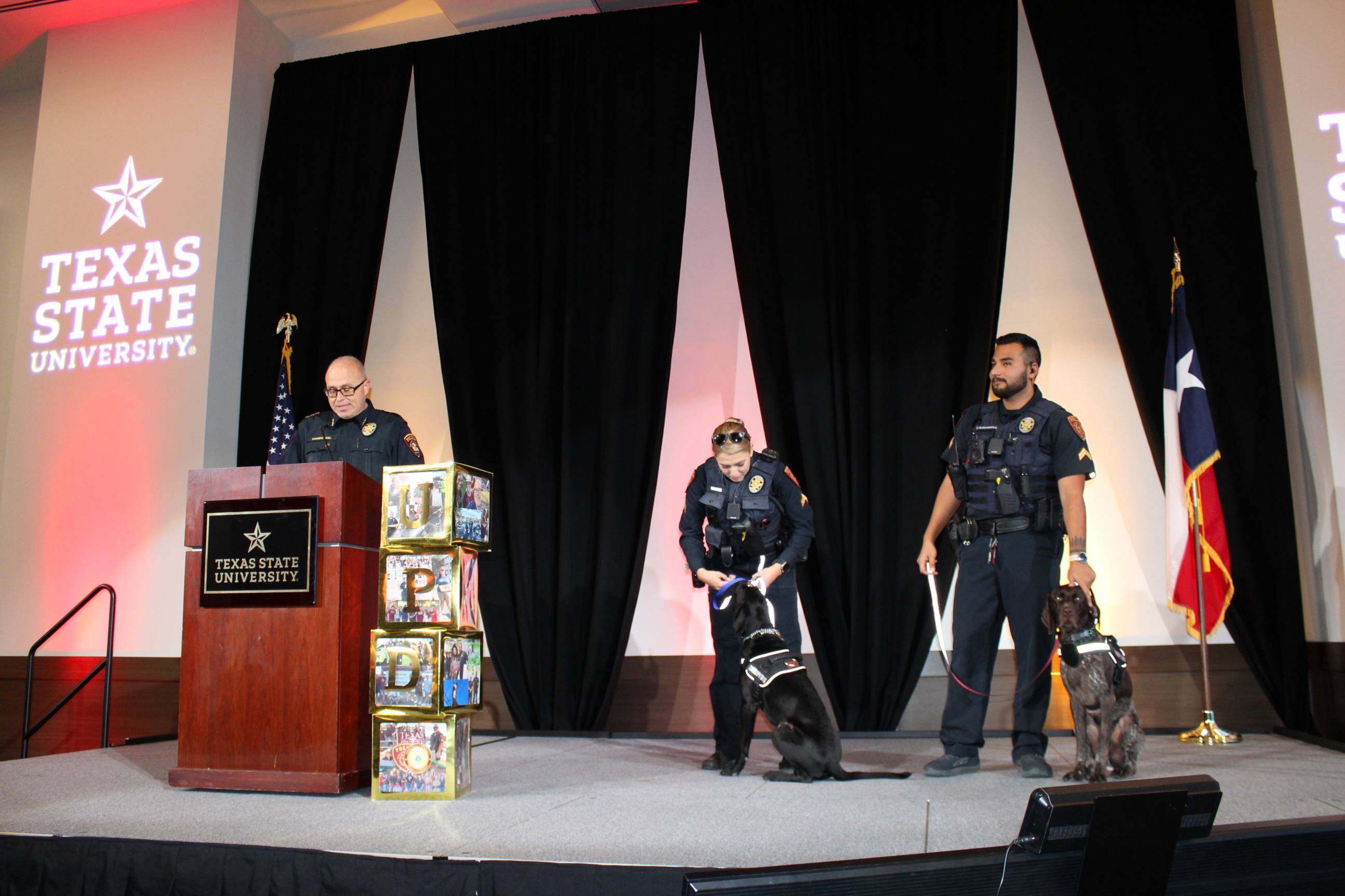 police chief gives speech while therapy k9s and their handlers stand on stage in police attire