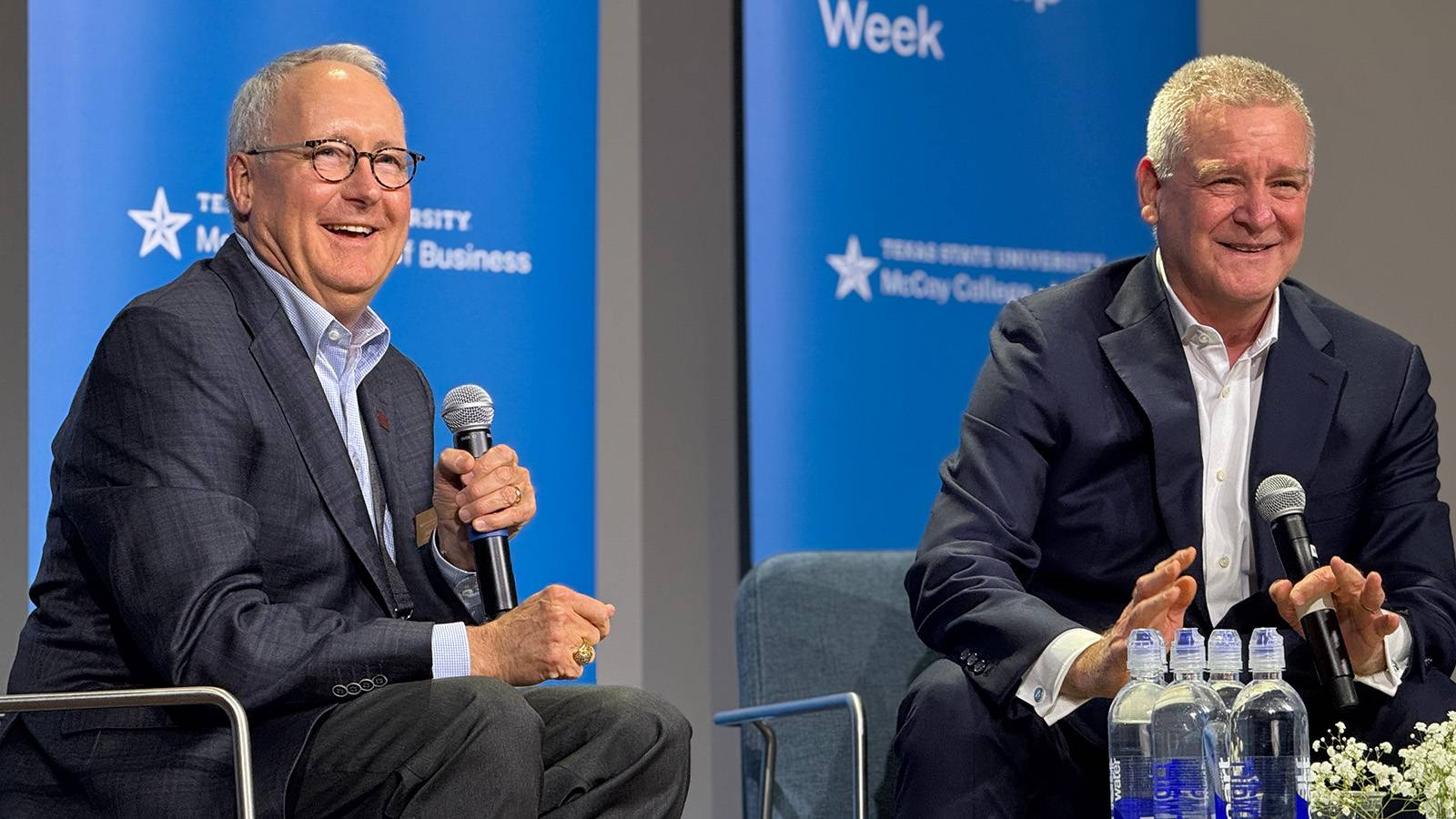two men in professional attire sit in chairs on a stage during a fireside chat