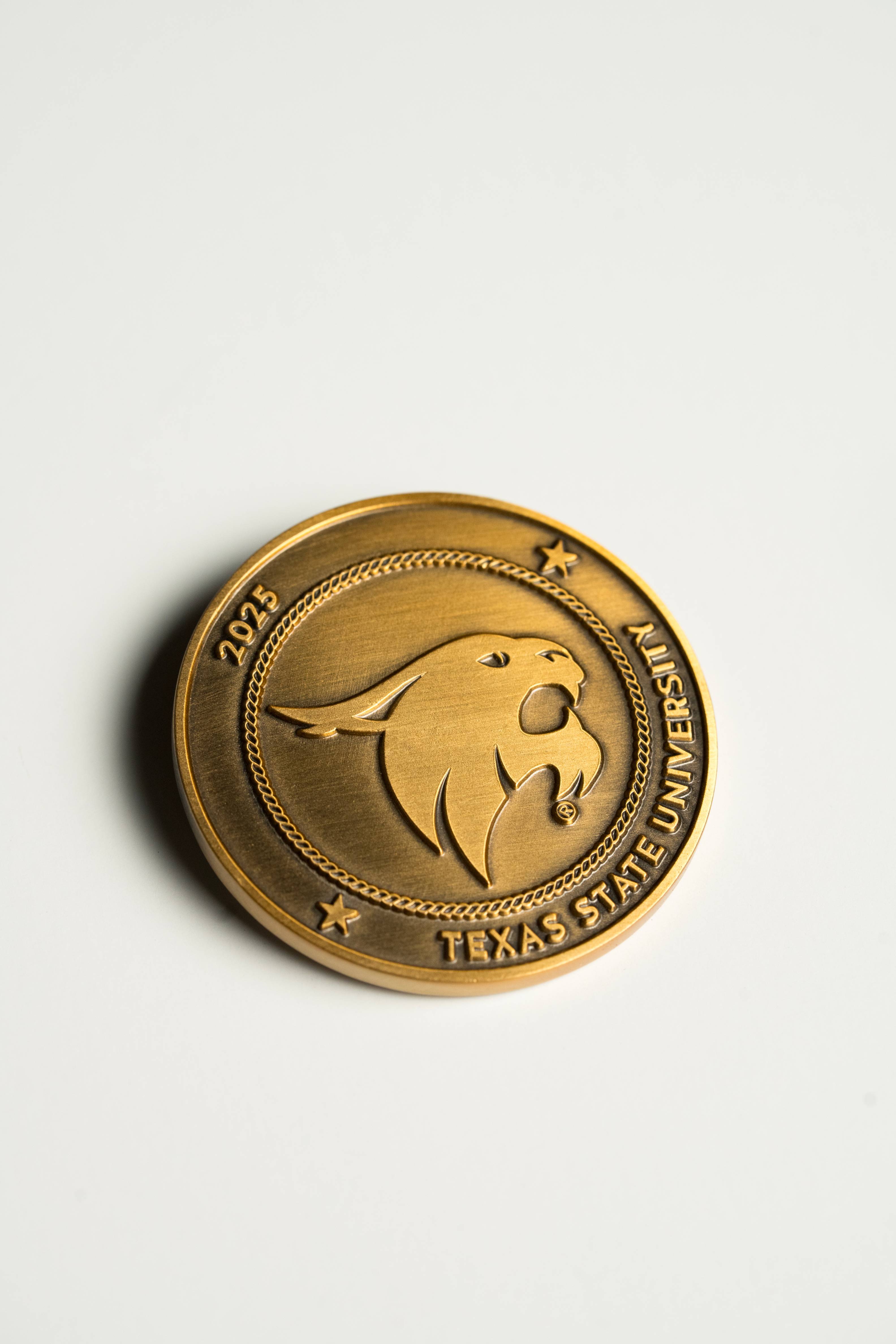 close up of a gold coin that reads "2025 Texas State University" with the university's SuperCat Logo in the center