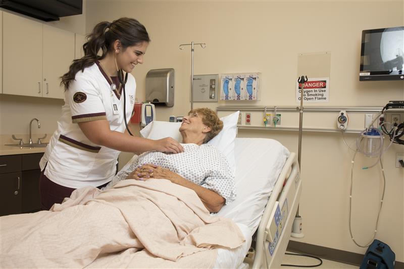 Smiling Texas State Nursing student holding a stethoscope on a smiling patient's chest as the patient lays down. 