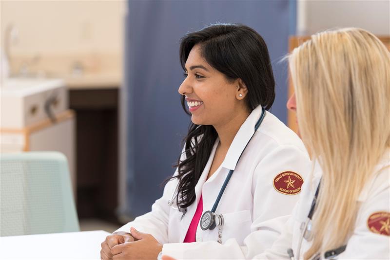 Two graduate Texas State Nursing students, one with black hair and one with blonde hair, smiling in class. 