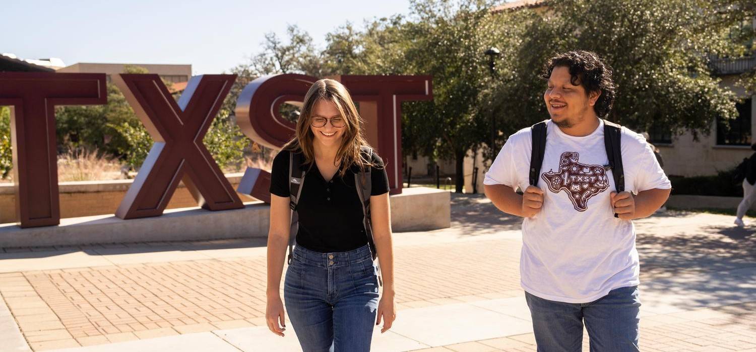 two students smile and talk while walking past TXST acronym sign on campus