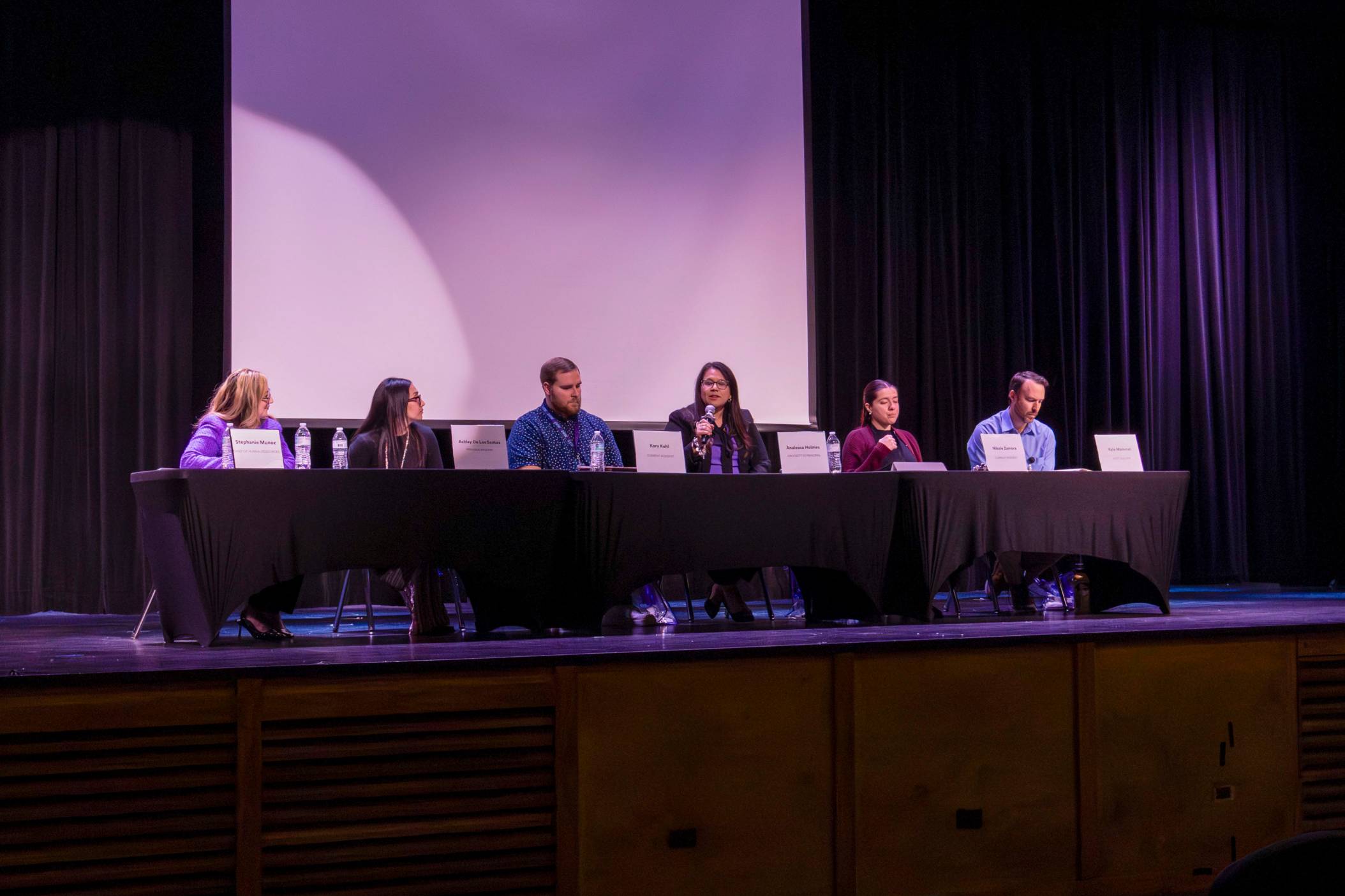 a panel of six individuals sit behind table on top of a stage