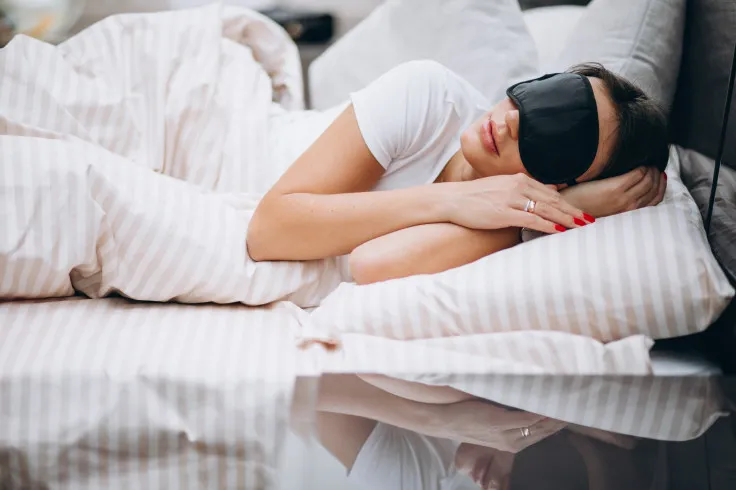 A woman wearing a black sleep mask sleeps in her bed.
