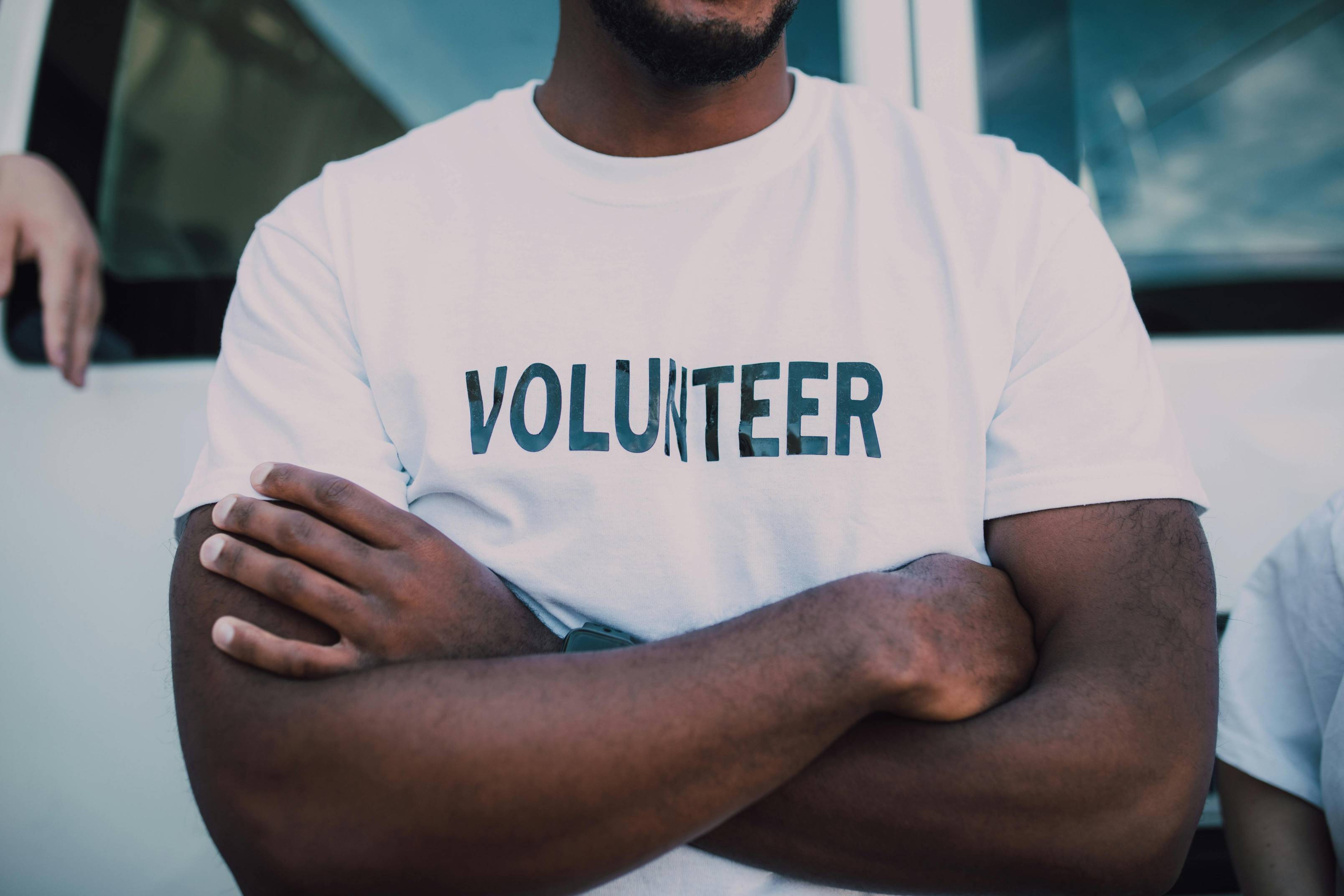 Close-up of a volunteer standing with arms crossed, wearing a white T-shirt labeled “VOLUNTEER.” The image represents community service, nonprofit organizations, volunteer engagement, and social impact, highlighting the mission-driven work involved in starting and managing a successful nonprofit.