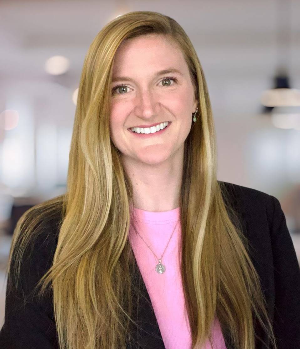 Blonde woman with pink shirt and black blazer smiling in front of blurred office background