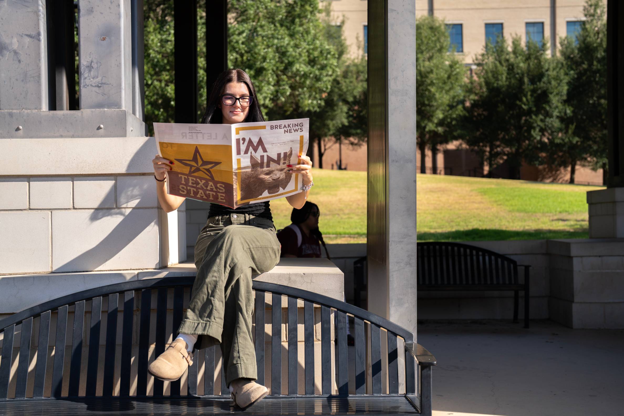 woman sitting on top of a metal bench with legs crossed. she is reading a newspaper reading 'I'm in!"