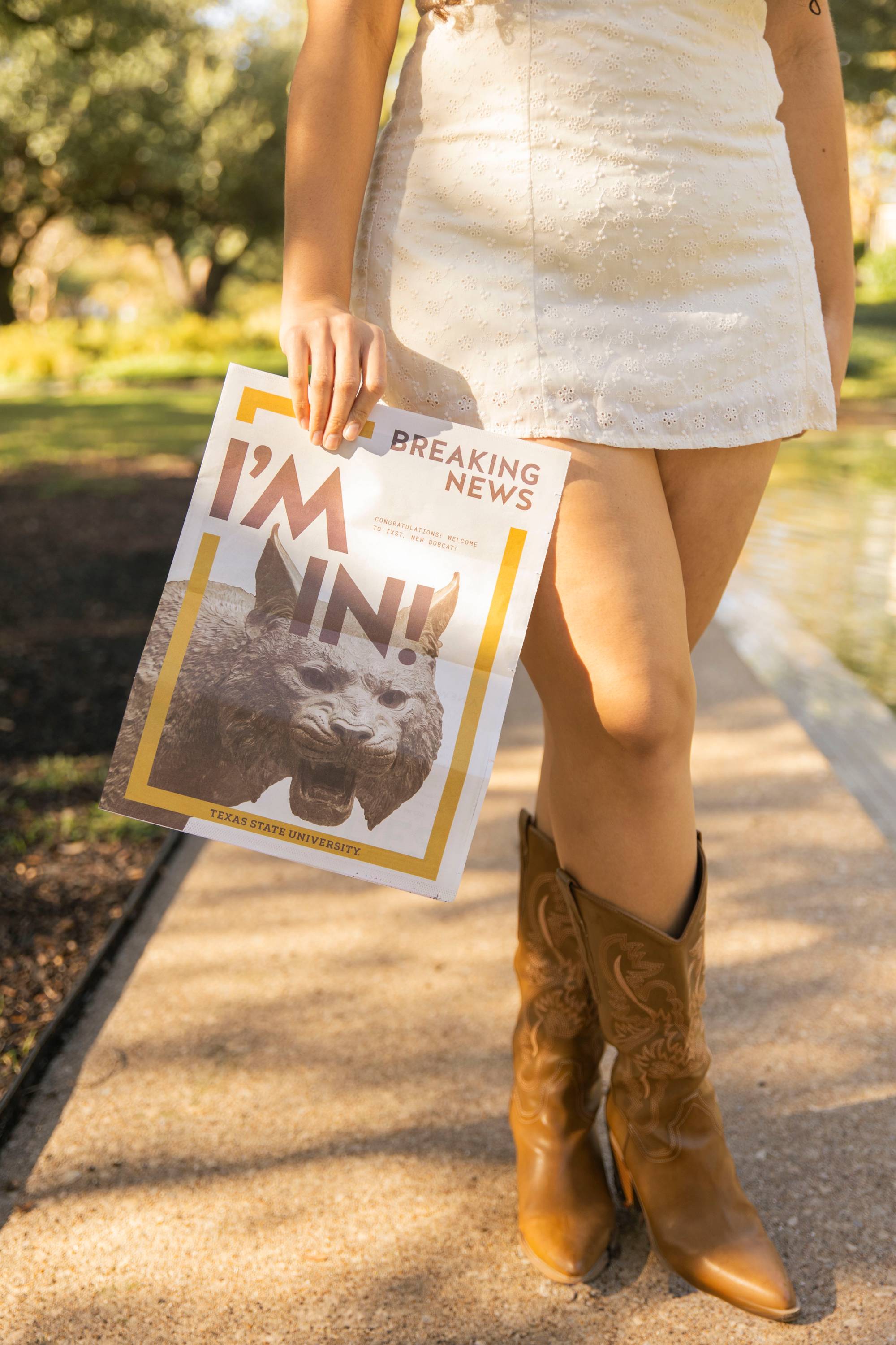 close up of woman holding a newspaper reading "Breaking New I'm in!" next to her legs. she is wearing a white dress and brown cowboy boots. 