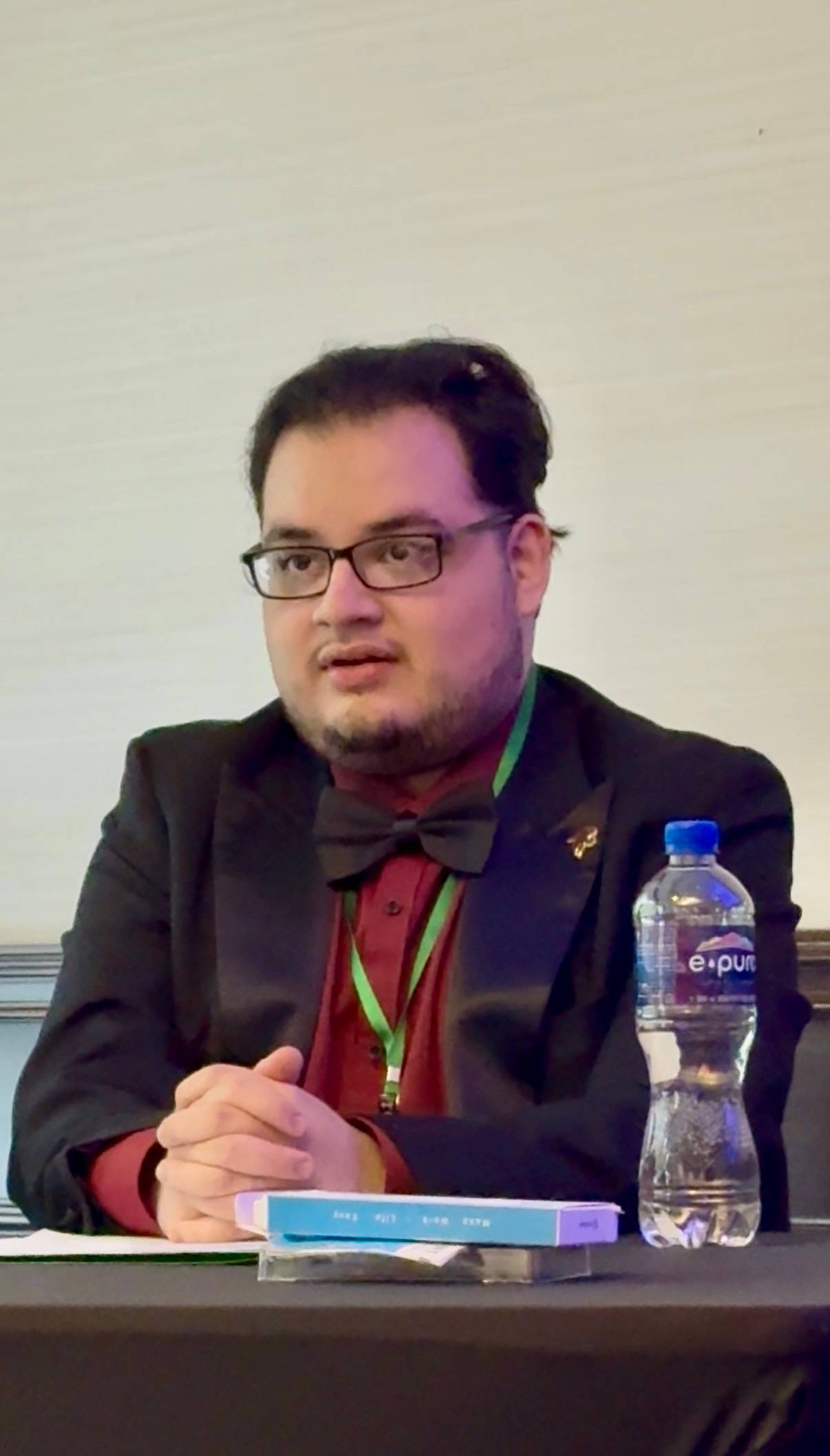 man wearing suit with bow tie, glasses, sitting at table, bottled water