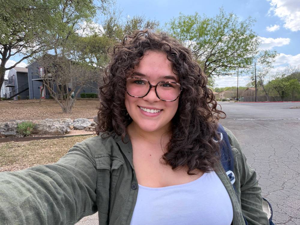 Vice President Elianna Knaggs of Sigma Tau Delta, a woman with curly hair and glasses smiles while taking a selfie. 