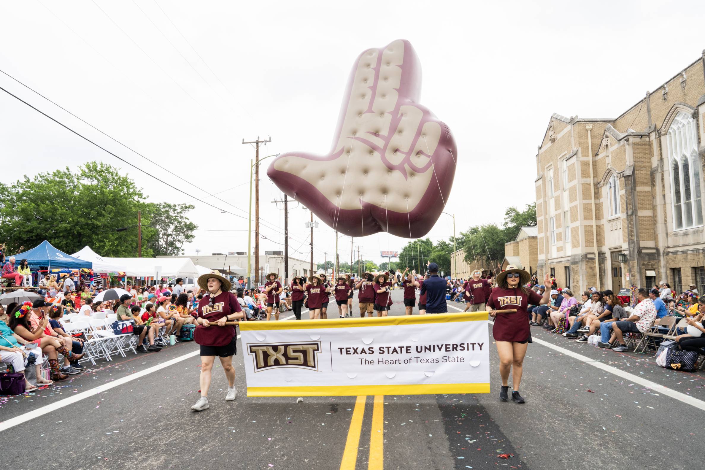 Texas State University students carry a TXST banner during the 2025 Fiesta San Antonio parade, with a large "Heart of Texas" hand sign balloon floating above and crowds lining the street.