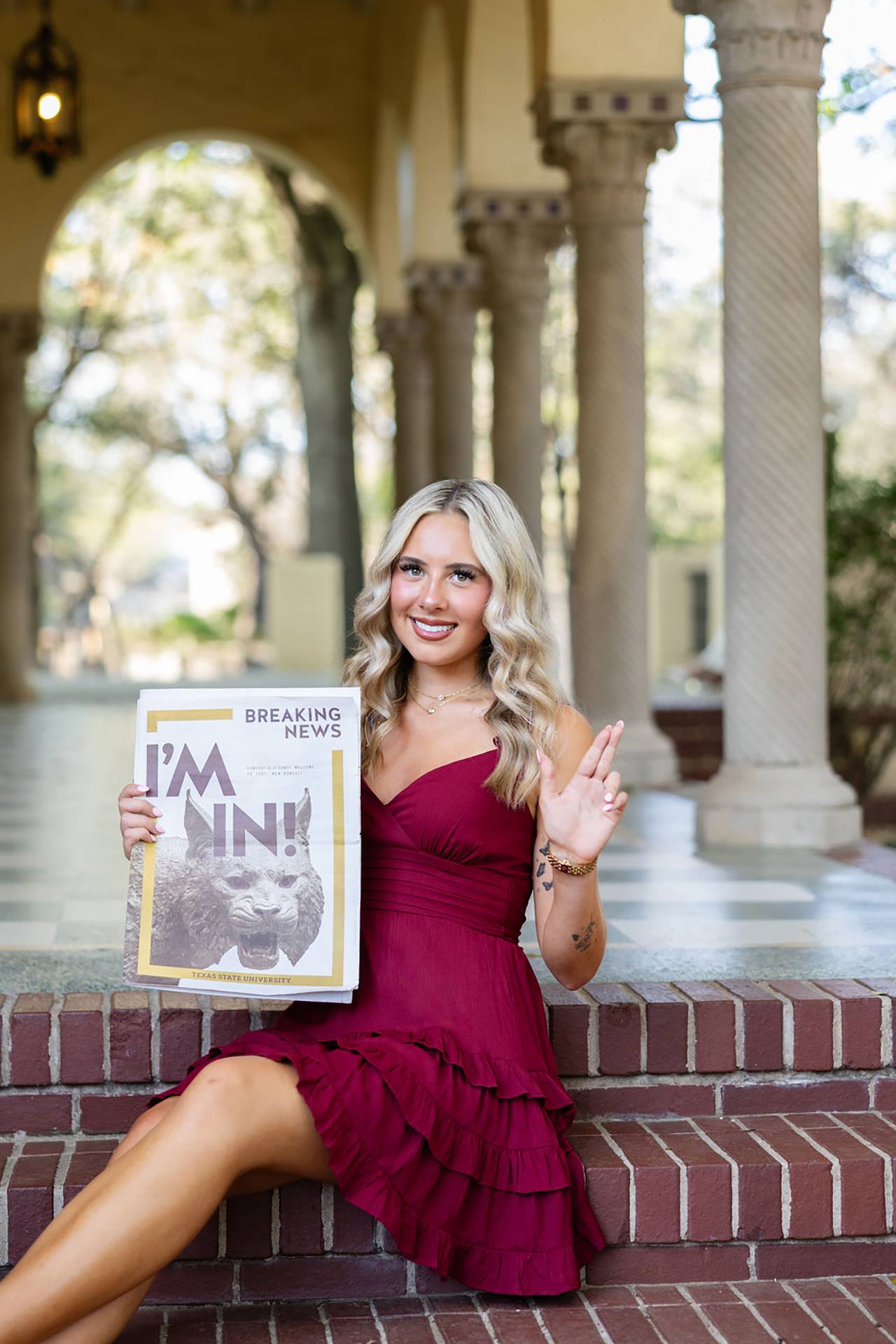 woman in maroon dress sitting on brick stairs while holding up the txst hand sign with her left hand and a newspaper reading "I'm in!" in her right hand.