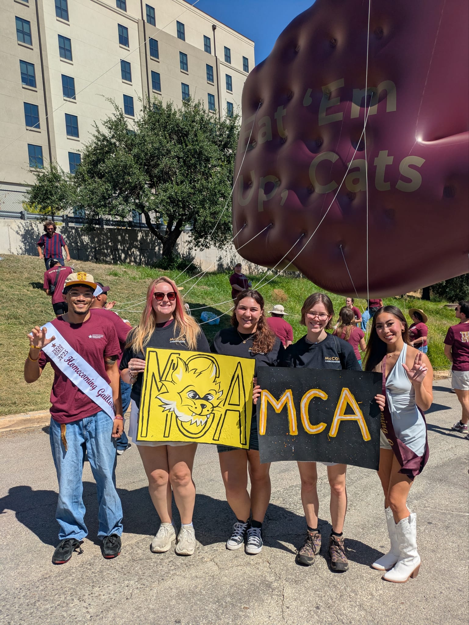 Five McCoy Ambassadors walking in the Homecoming Parade, holding two signs and a Texas hand sign balloon