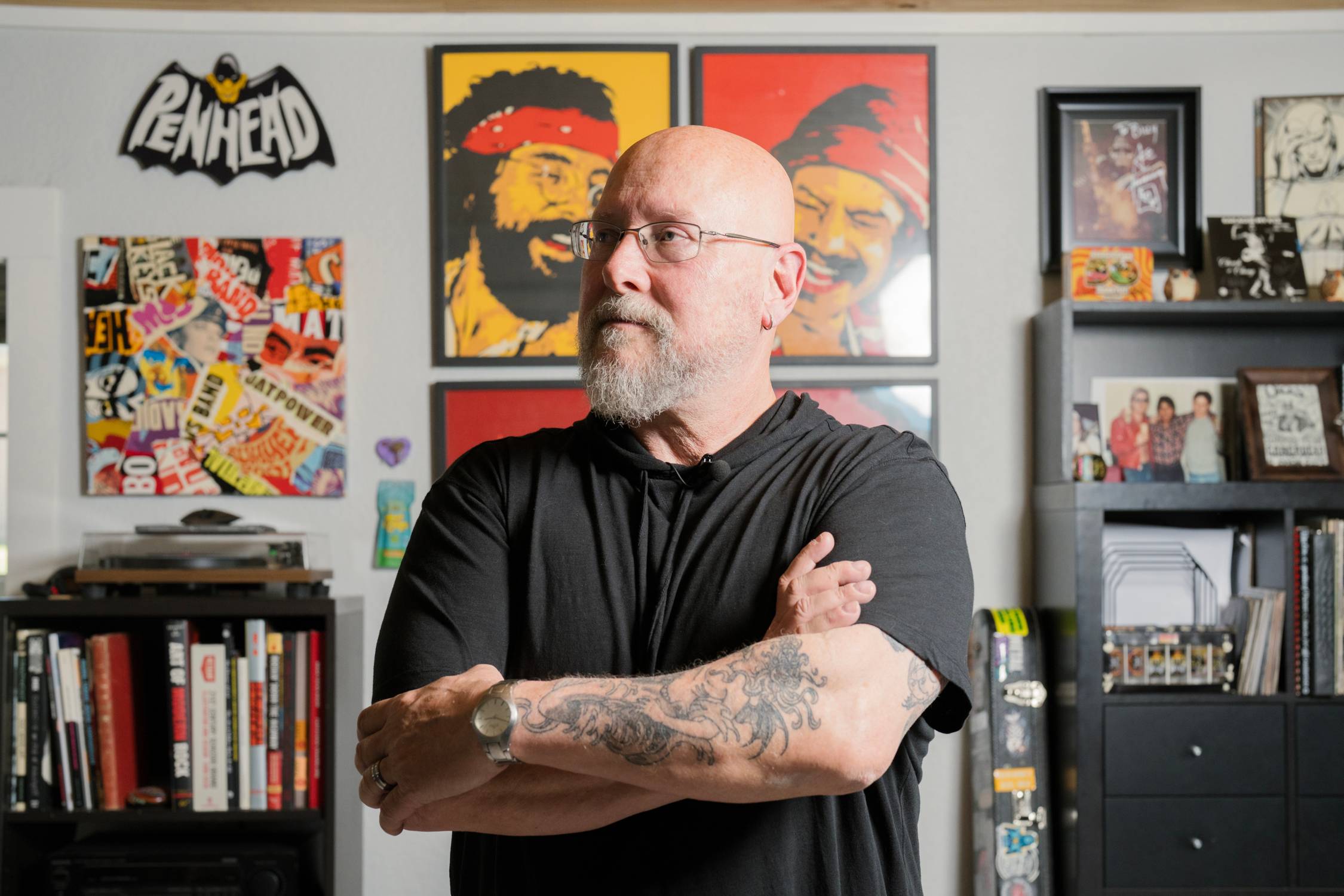 Older man standing with arms crossed with colorful portraits on the wall behind him. 