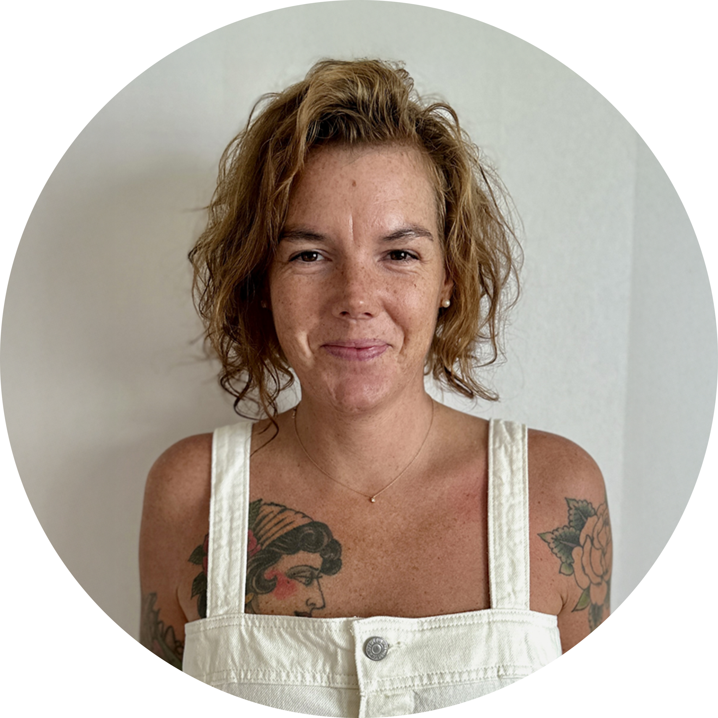 Swedish woman with short curly hair, tattoos, and white overalls.