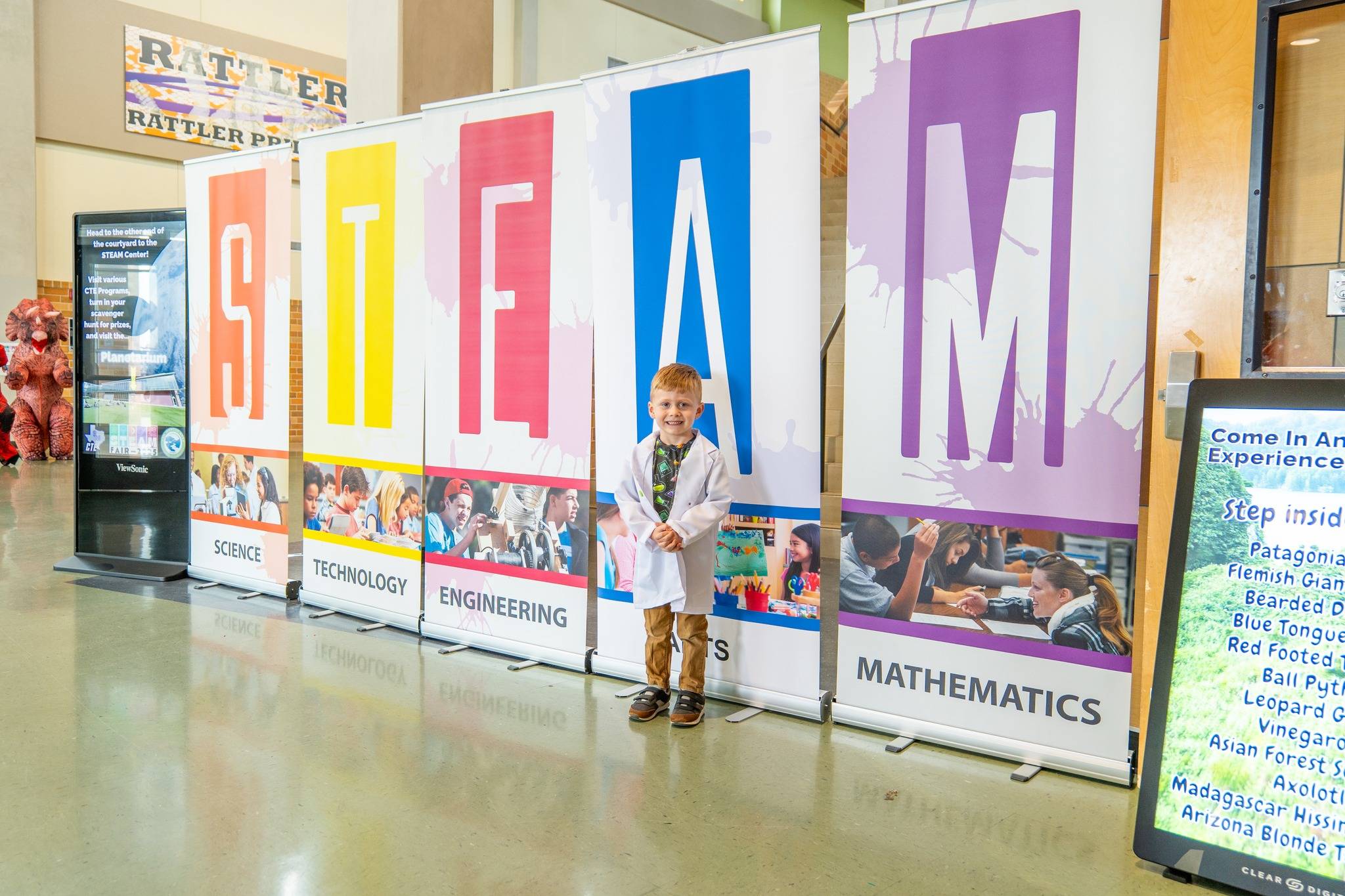 young child in white lab coat standing in front of large, colorful sign that reads STEAM