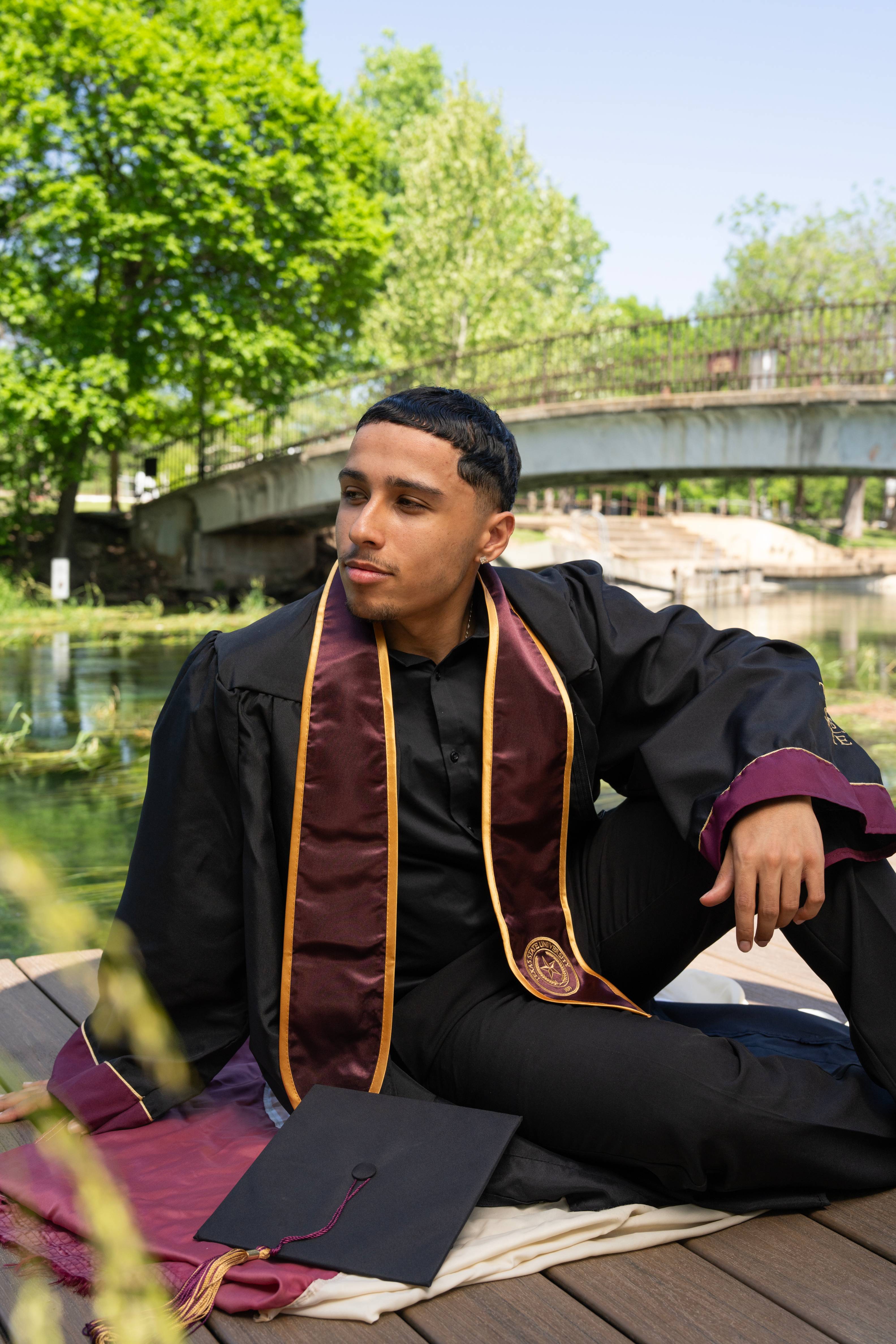 Graduate in cap and gown sits on a ledge at Sewell Park, looking off to the side with the San Marcos River and a bridge visible behind him.