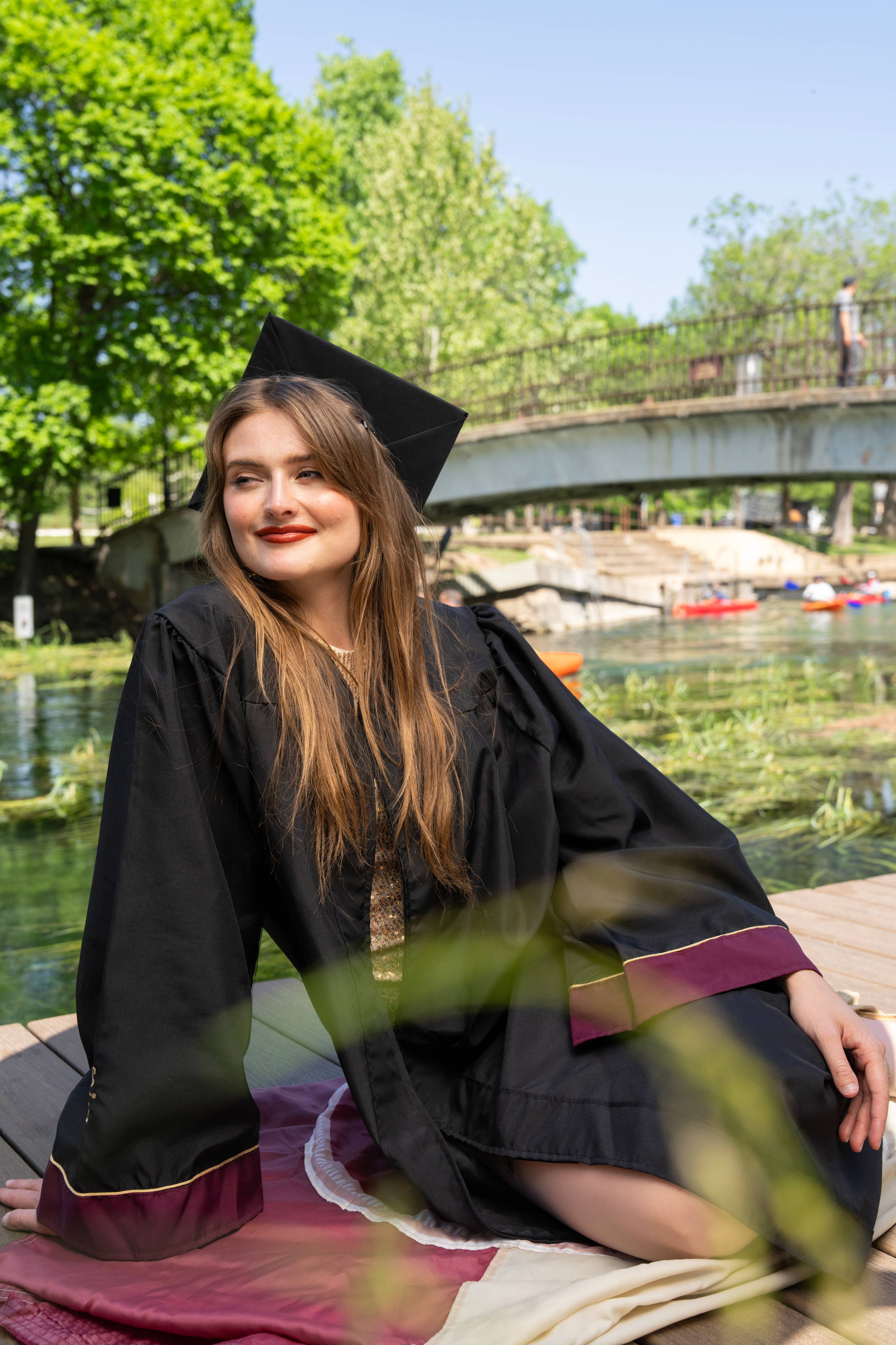 Graduate in cap and gown smiles at the camera at Sewell Park, with the San Marcos River, a bridge, and colorful kayakers visible in the background.