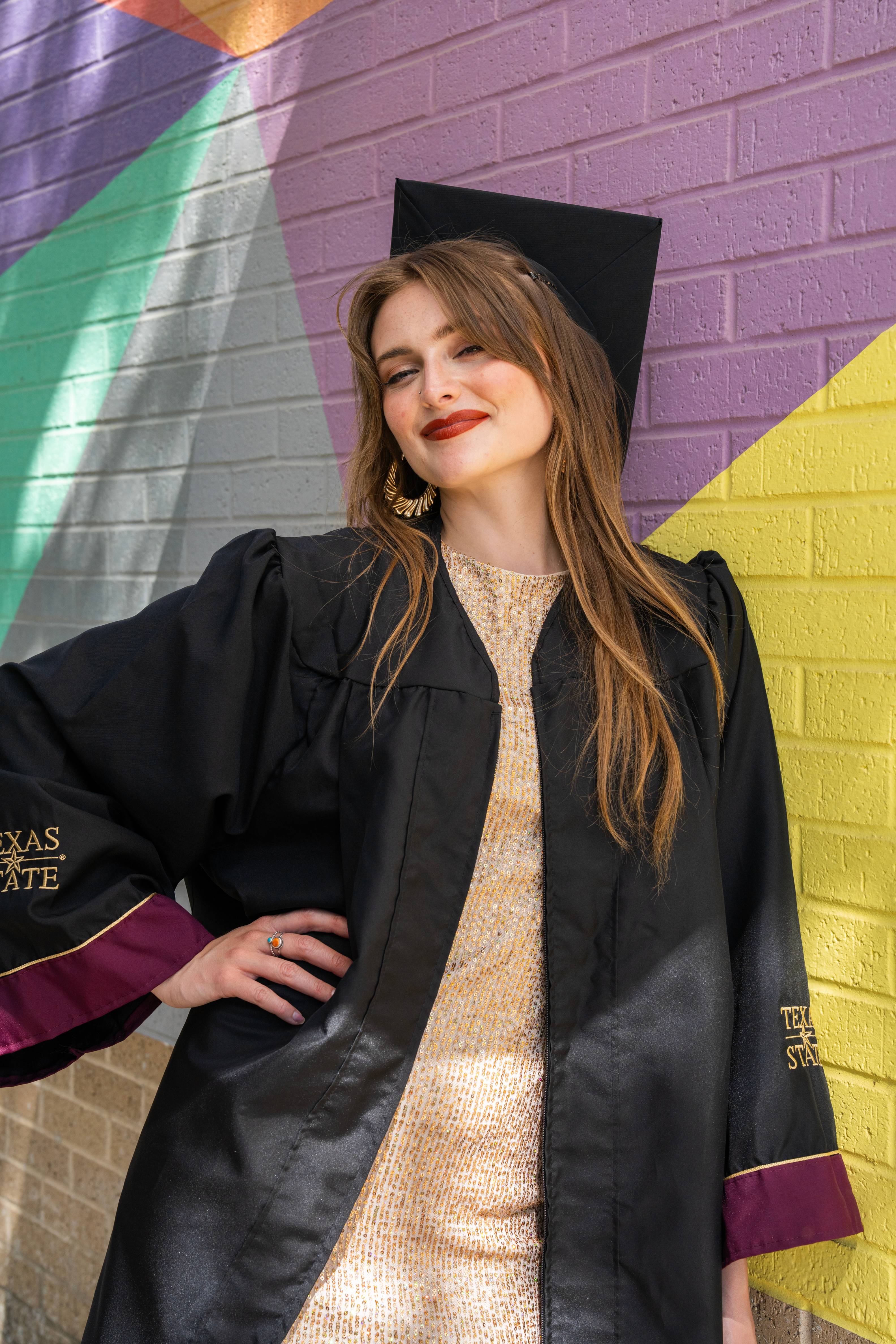 Graduate in a gold sequin dress and graduation gown poses confidently in front of the colorful One of a Kind mural.