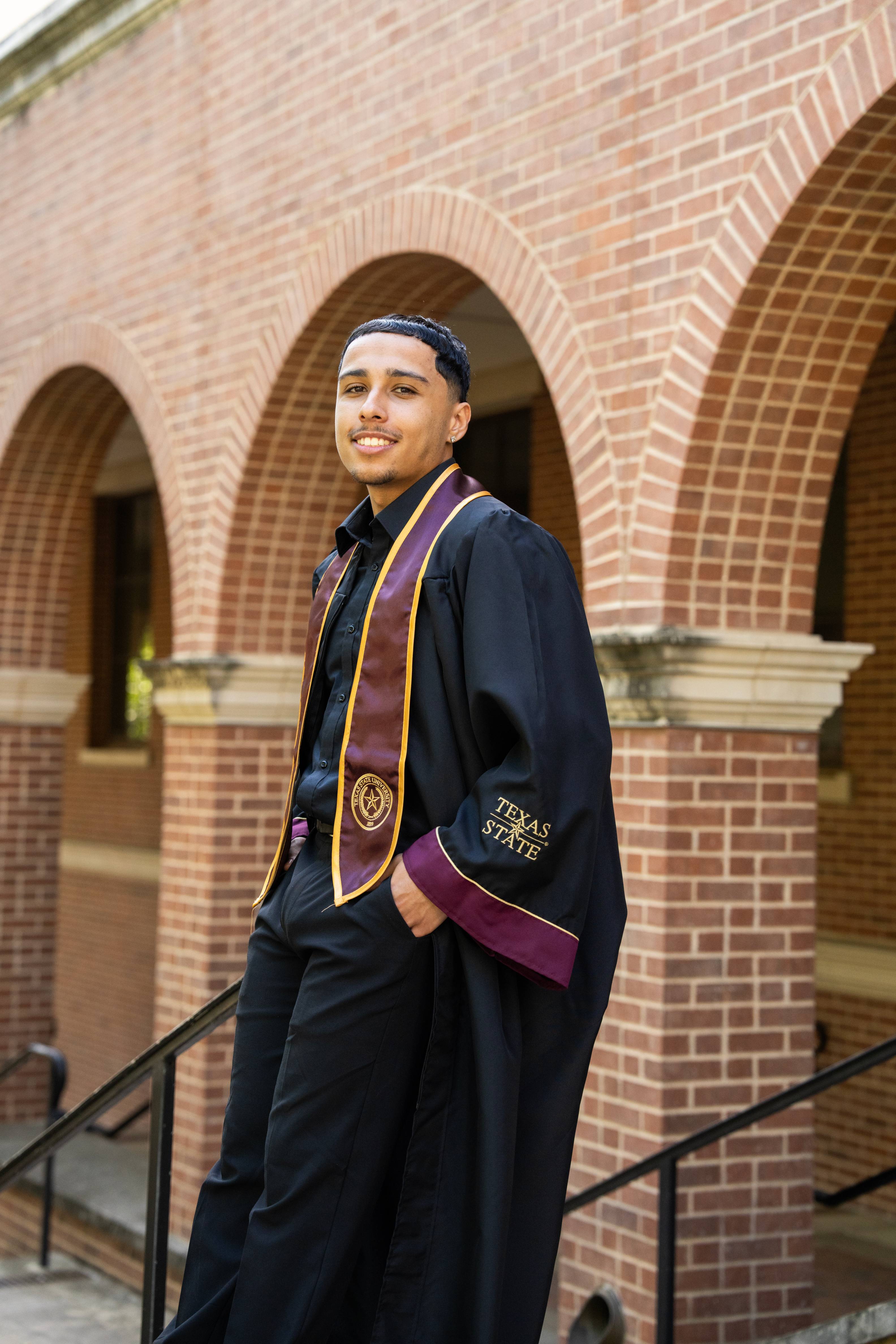 man in graduation regalia stands in front of multiple brick arches