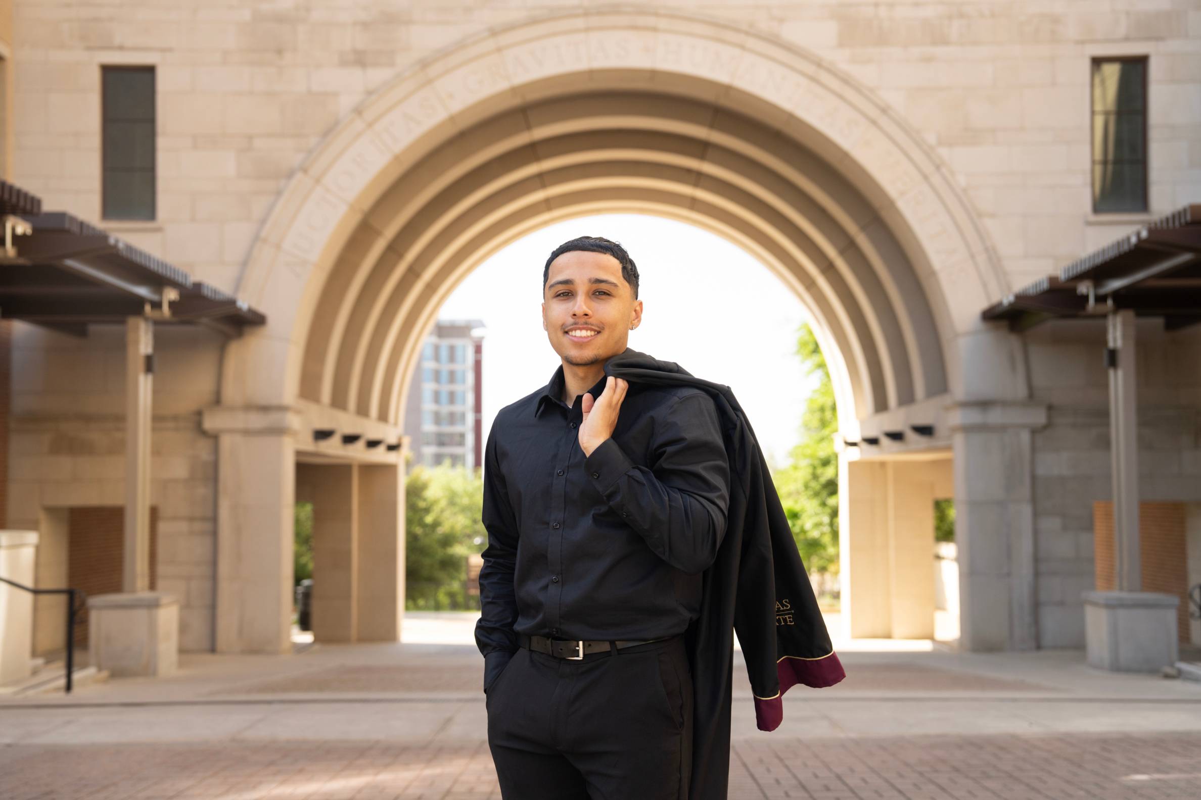 Graduate smiles at the camera with his robe over his shoulder under the UAC arch