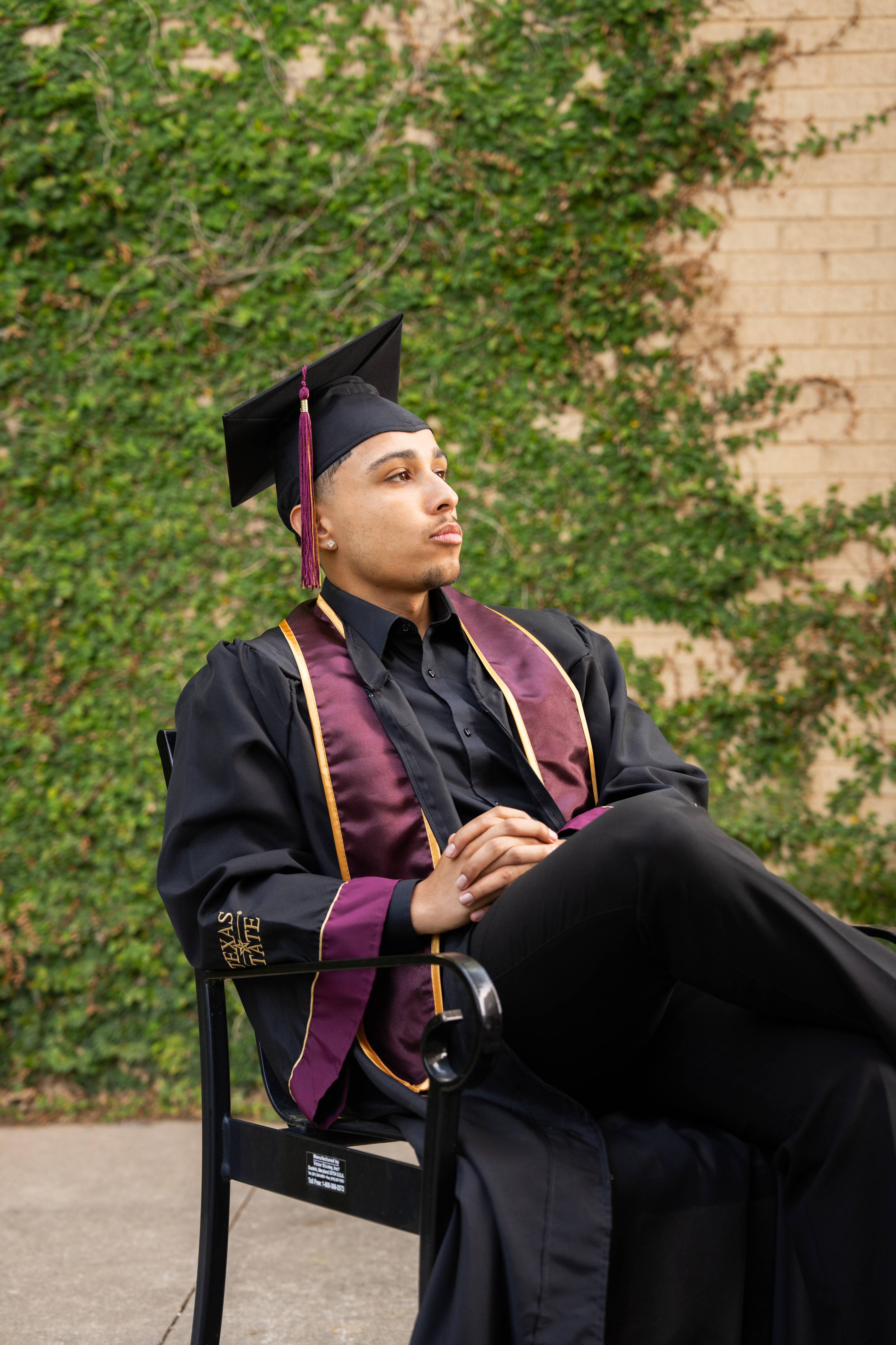 man in graduation regalia sits in front of brick wall covered in vines