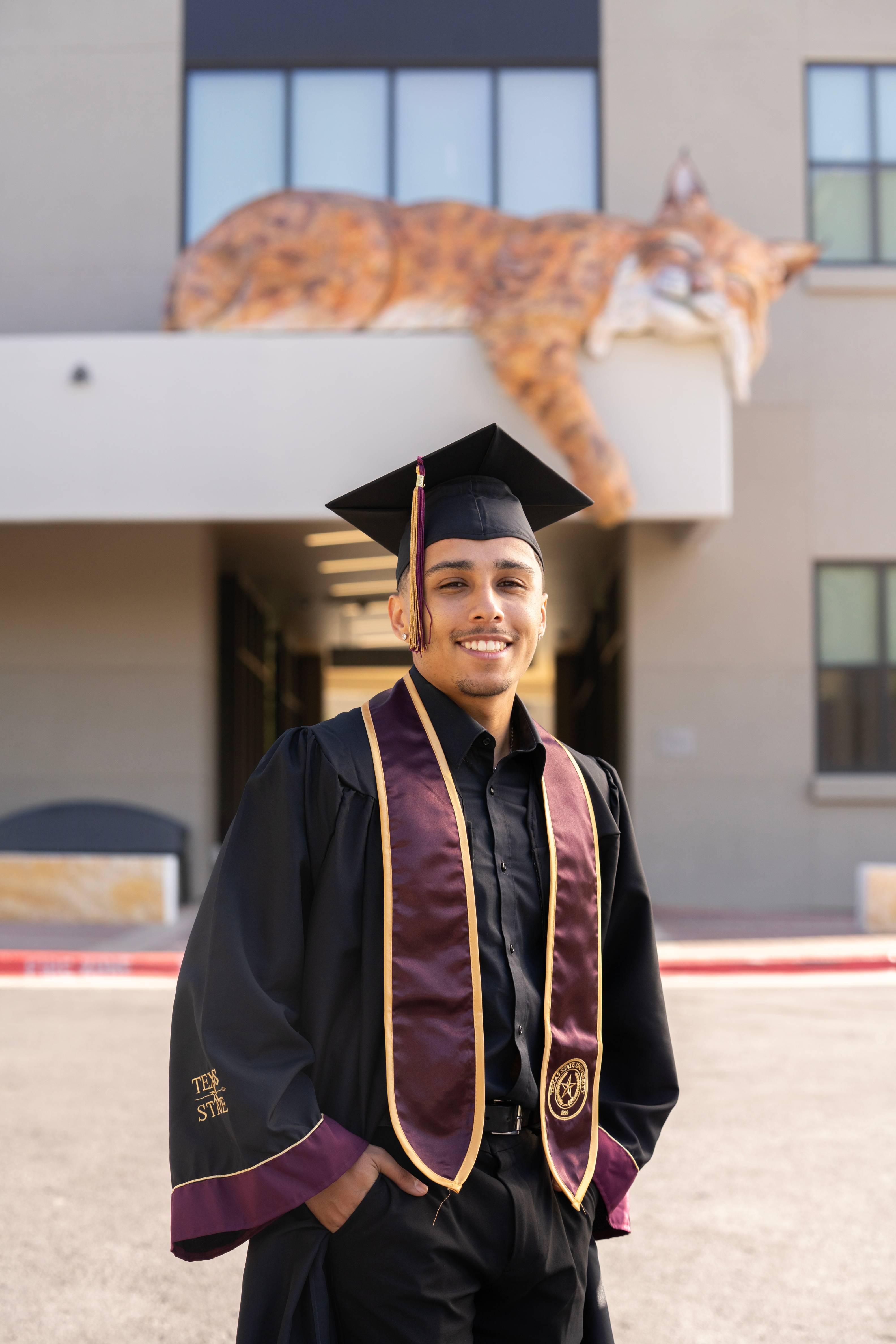 Graduate smiles at the camera in front of Cibolo Hall, with a painted bobcat sculpture visible on the building ledge behind him.