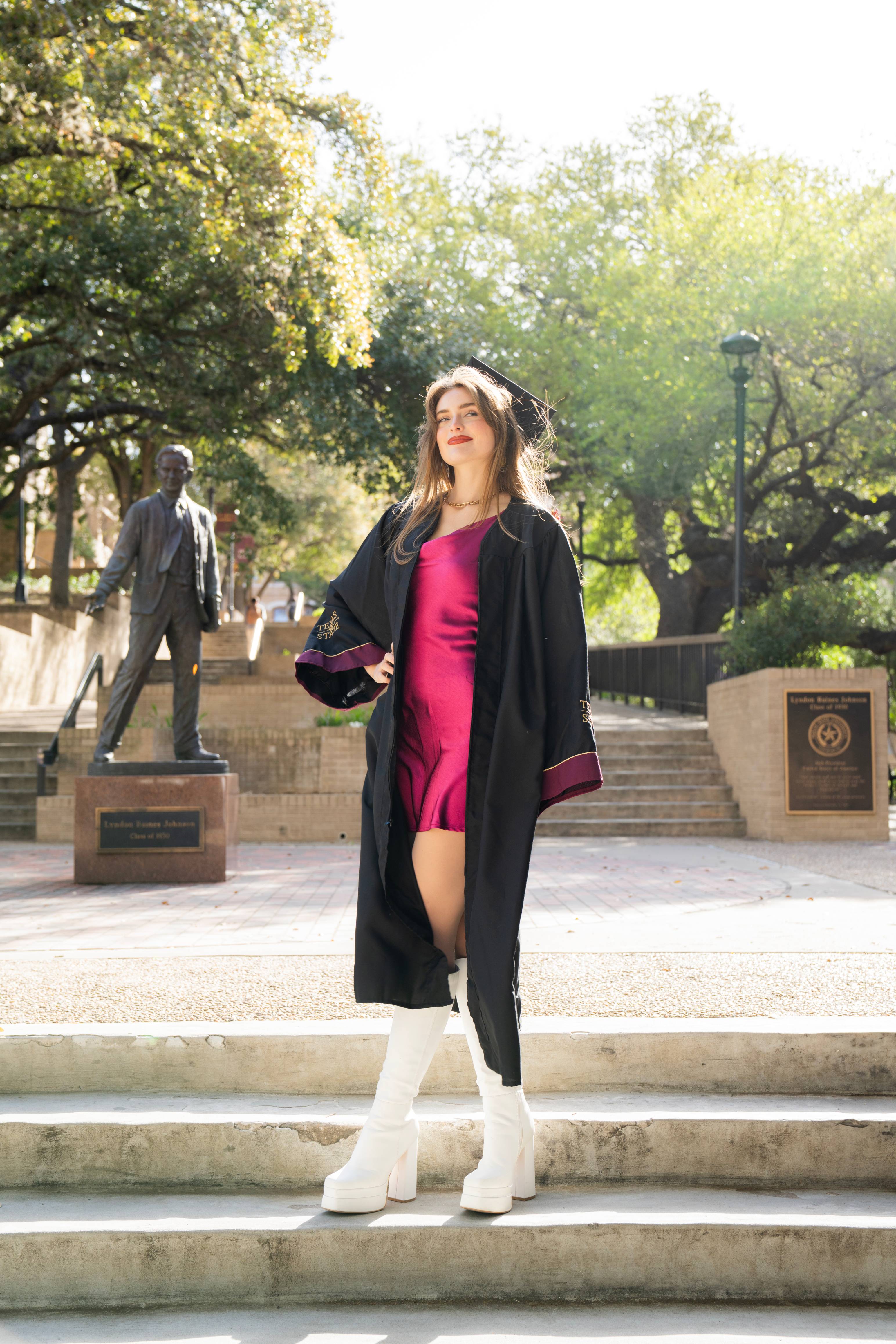 graduate in a pink dress and graduation gown poses in front of the LBJ statue, looking off to the right