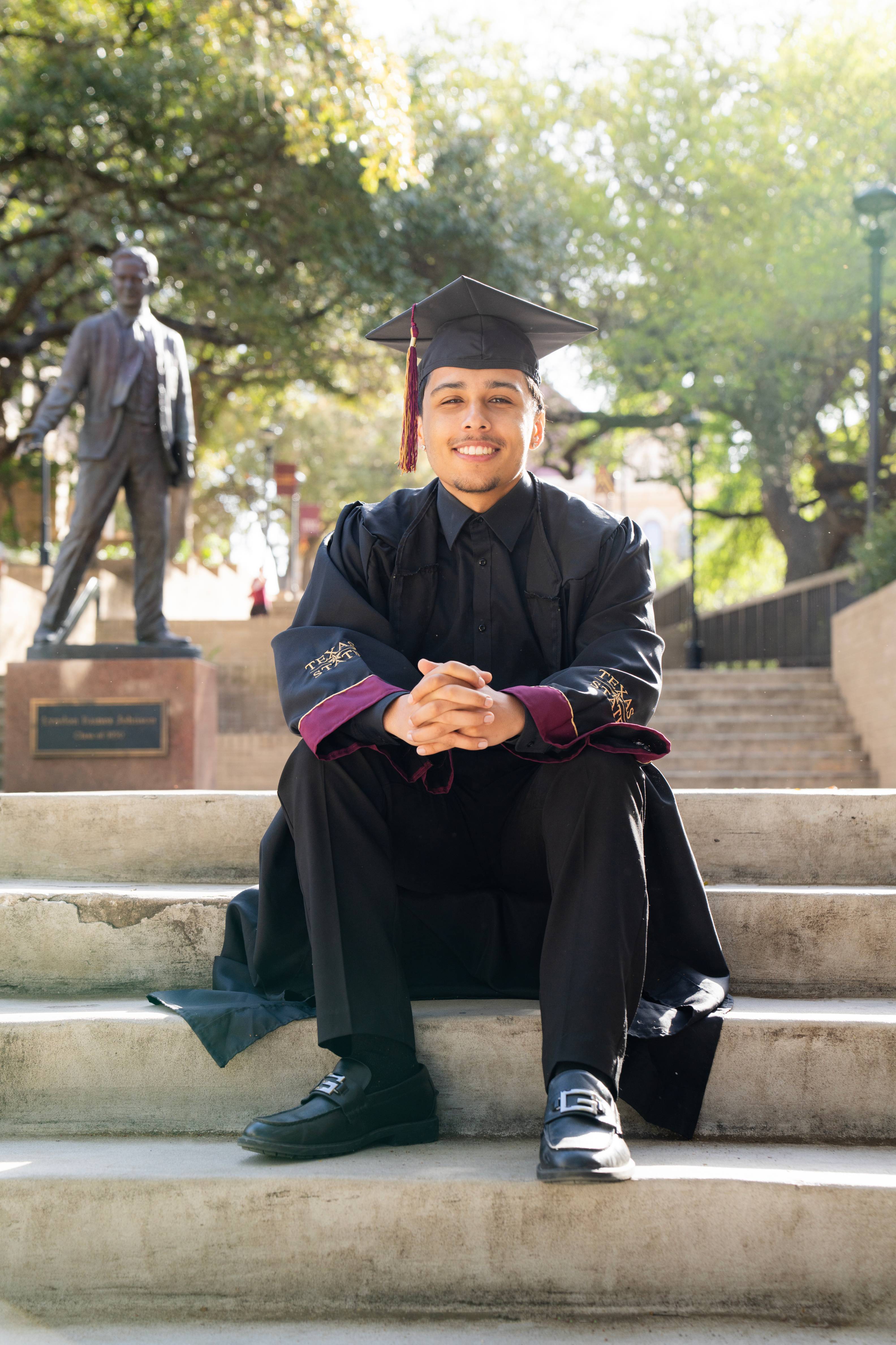 Graduate sits on the steps in front of the Lyndon B. Johnson statue on campus, smiling with his hands clasped.