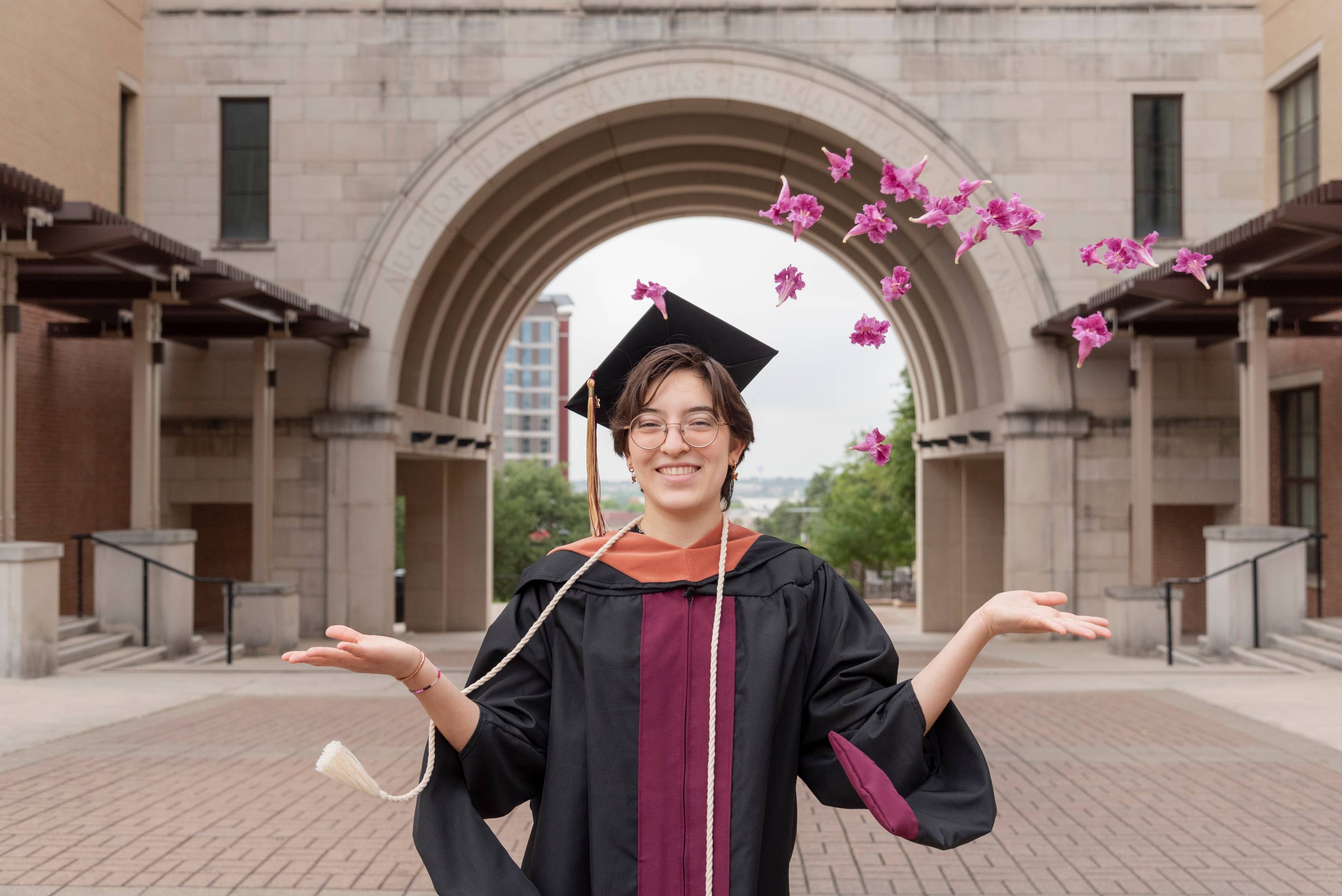 A graduate throws flower petals in the air, smiling at the camera under the UAC arch