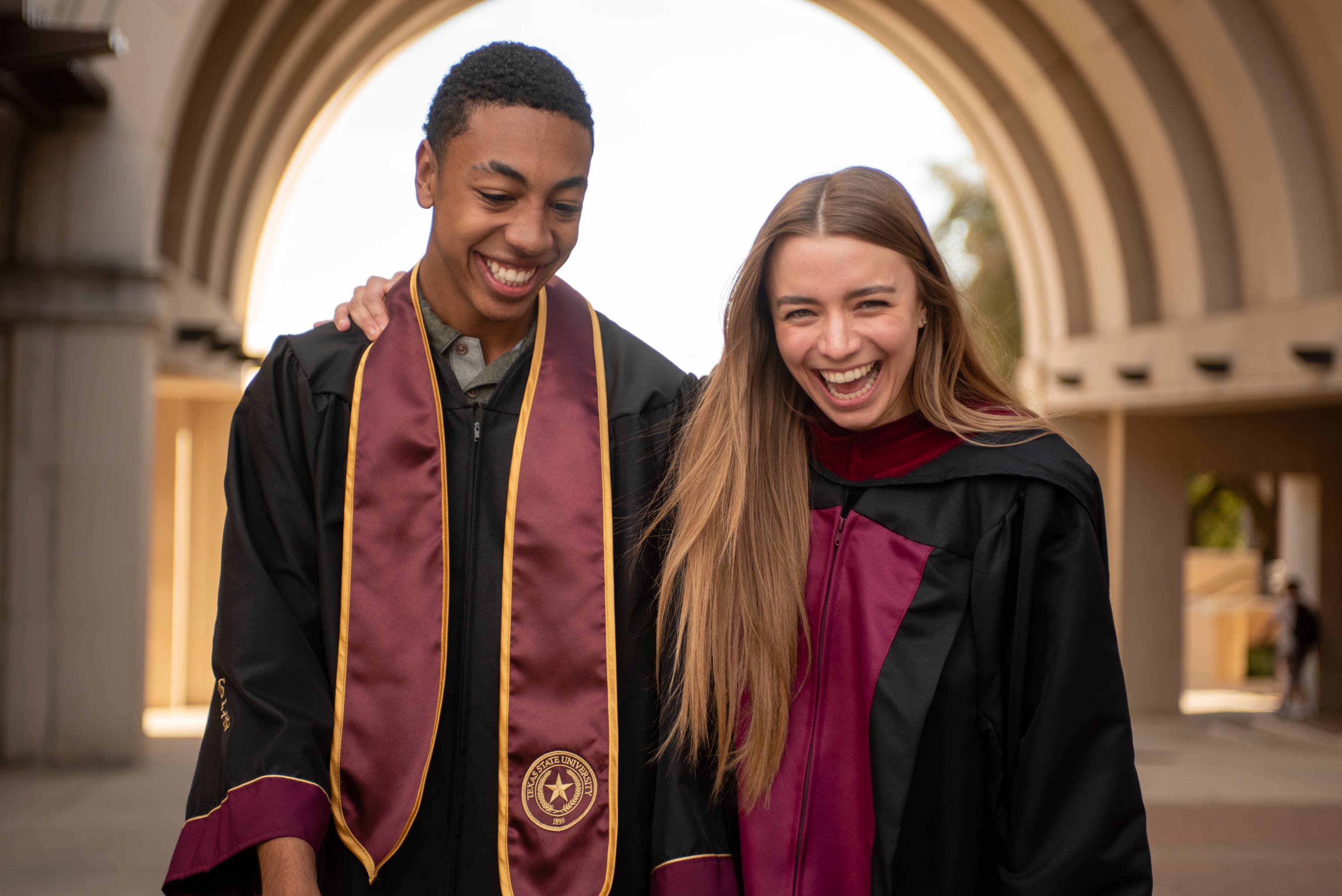 2 graduates laugh together under the UAC arch