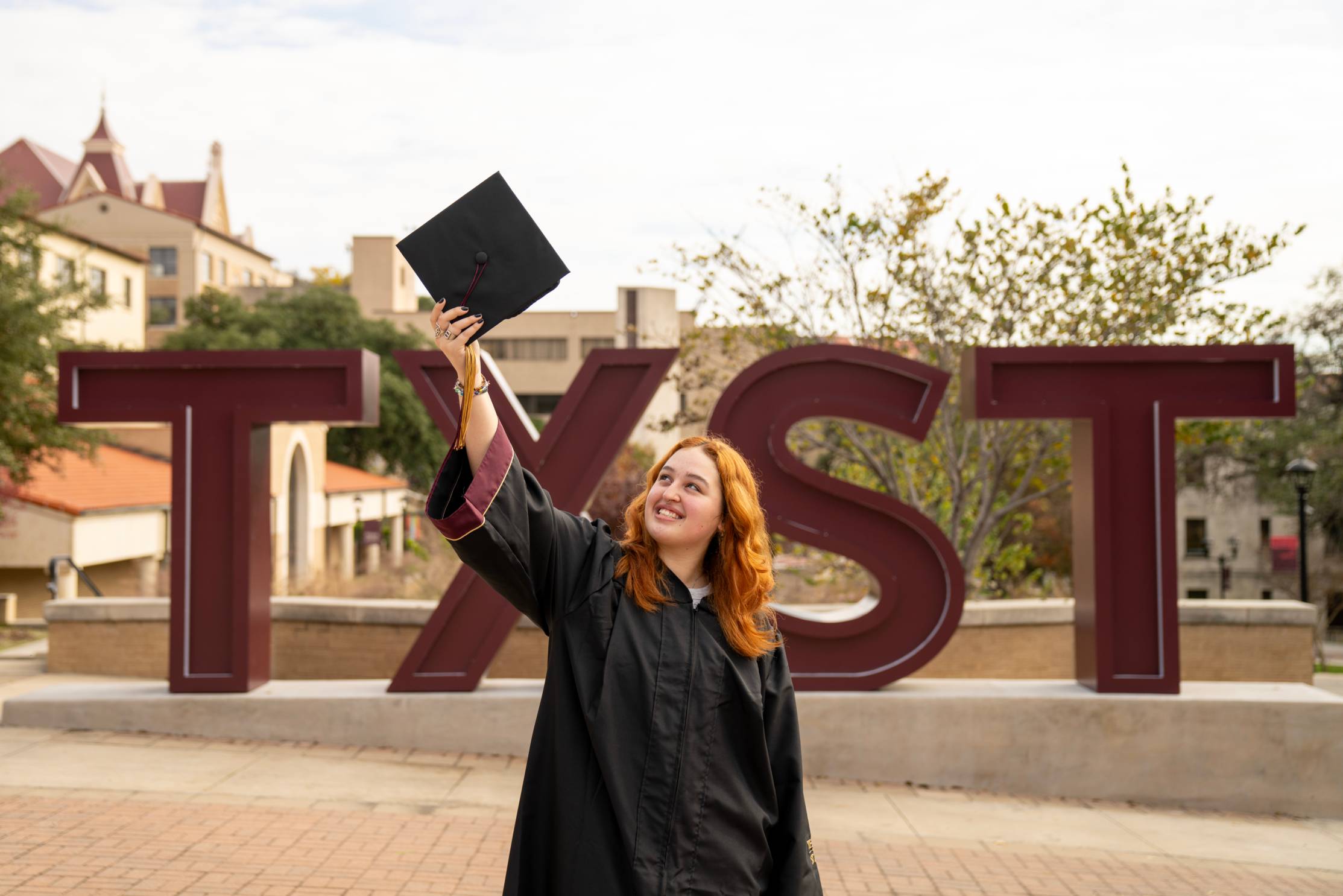 A graduate with red hair raises her cap toward the sky in front of the TXST sign on Bobcat Trail, smiling.