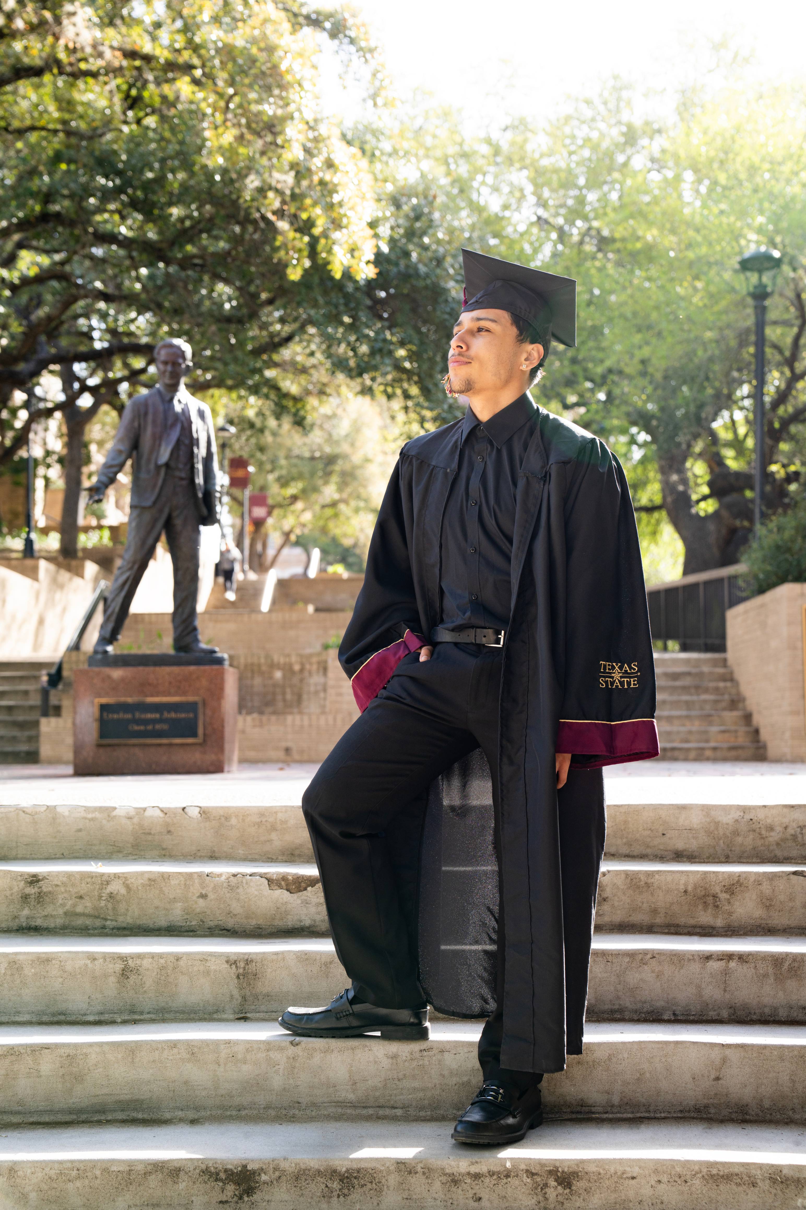 Graduate in his graduation gown poses in front of the LBJ statue, looking off to the left