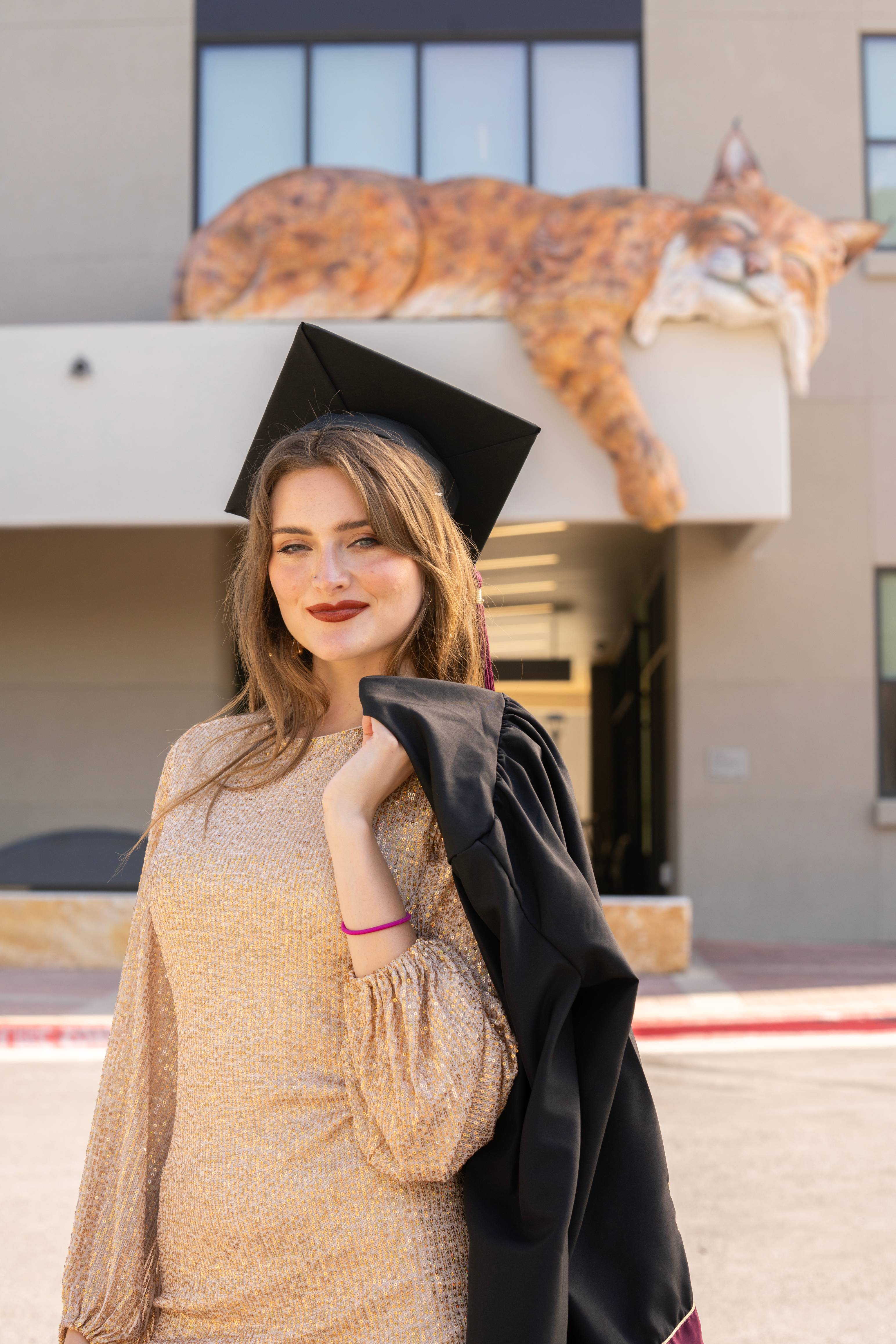 Graduate stands with her robe over her shoulder in front of Cibolo Hall, a painted bobcat sculpture visible on the building ledge behind her.