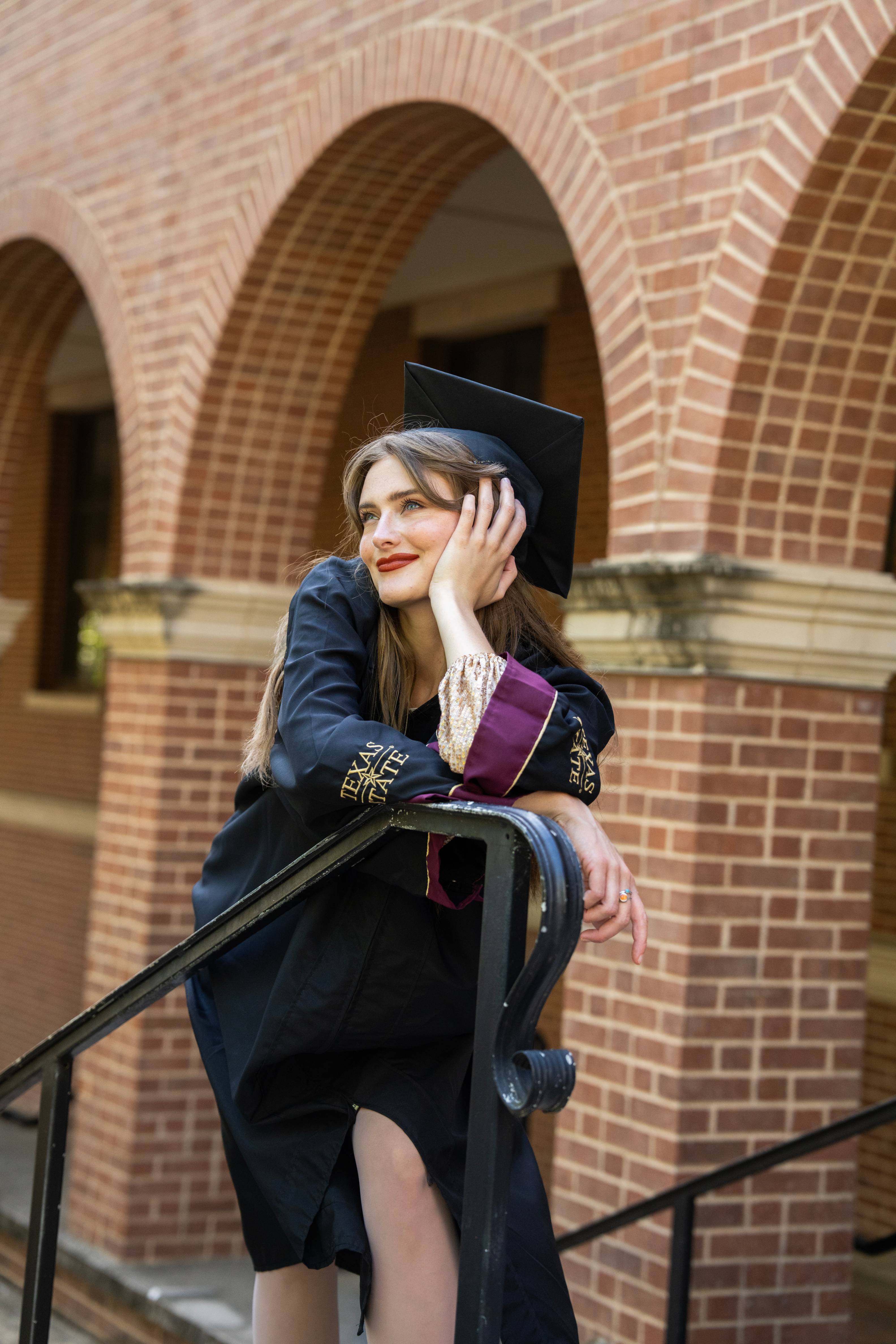 woman in graduation regalia leans on stair railing in front of multiple brick arches