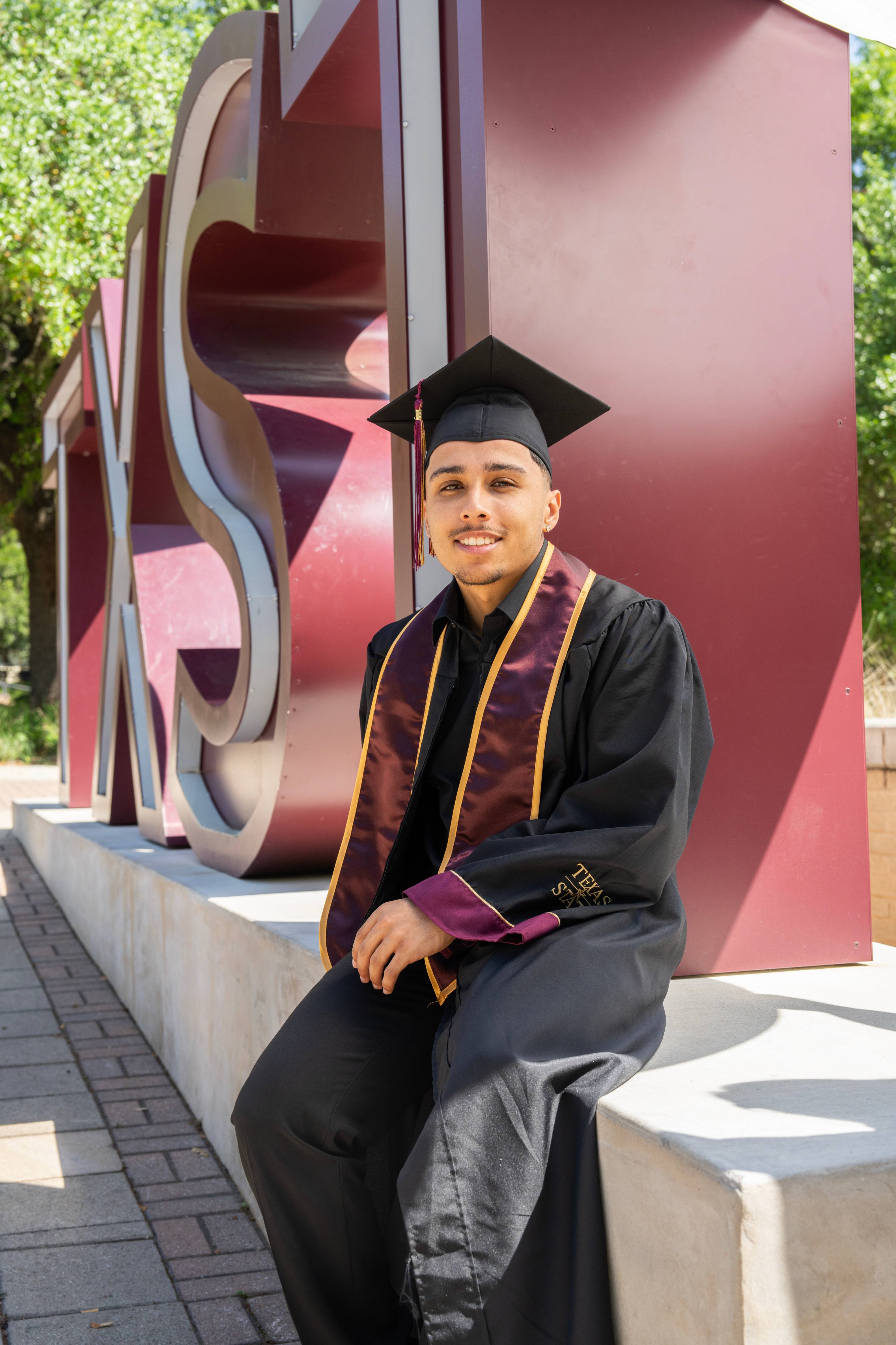 A sits against the base of the maroon TXST letters, wearing a maroon and gold stole, smiling at the camera. 