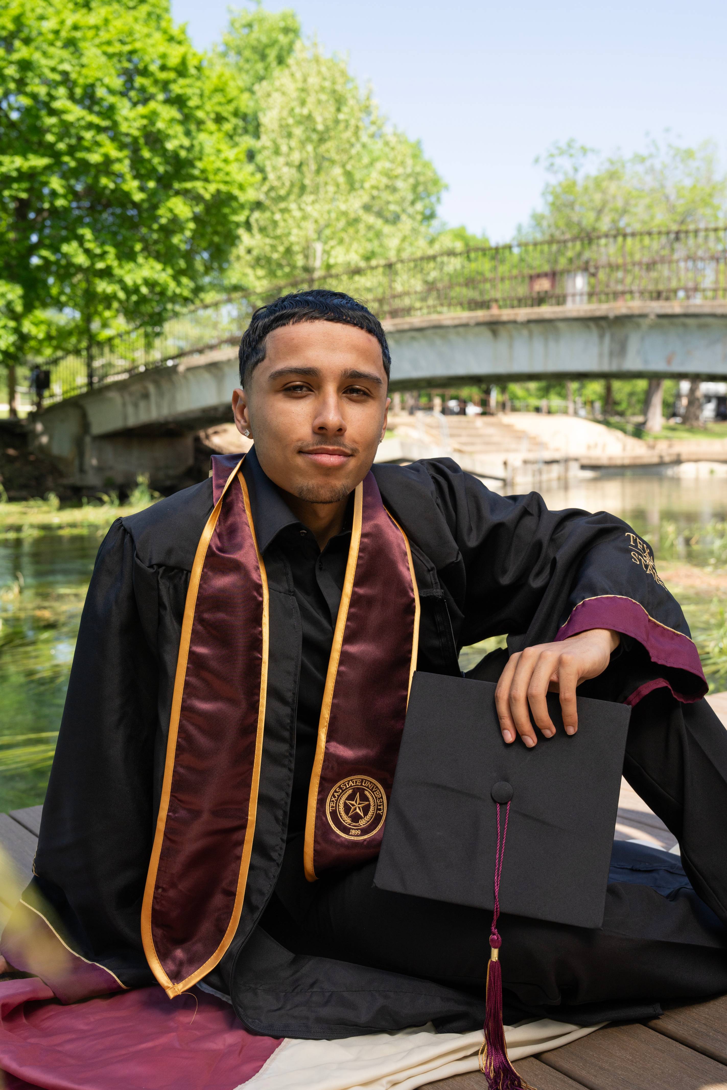 Graduate in cap and gown looks directly at the camera at Sewell Park, with the river and bridge softly blurred behind him.