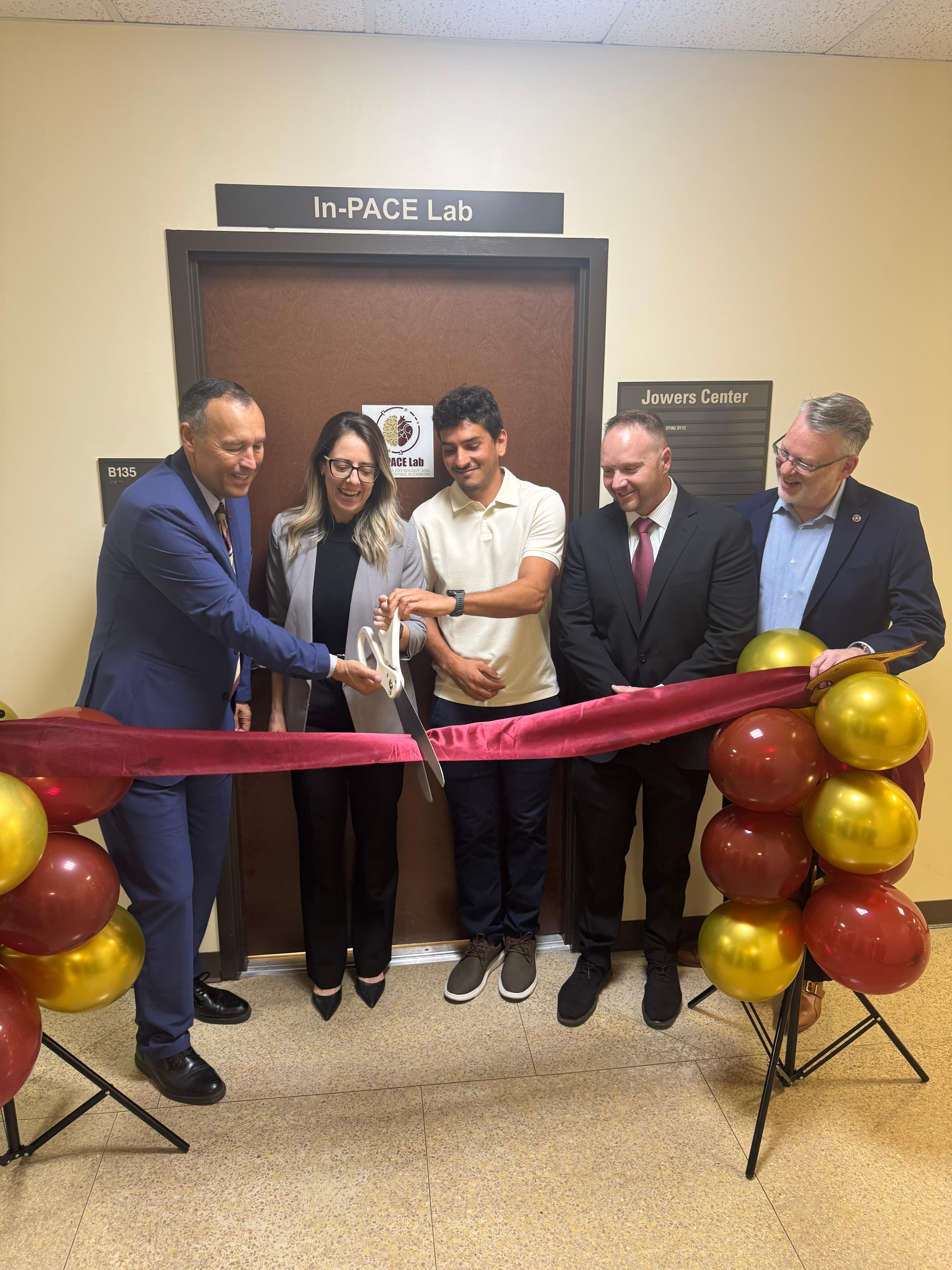 five people standing in front of a door cutting a large red ribbon with oversized scissors