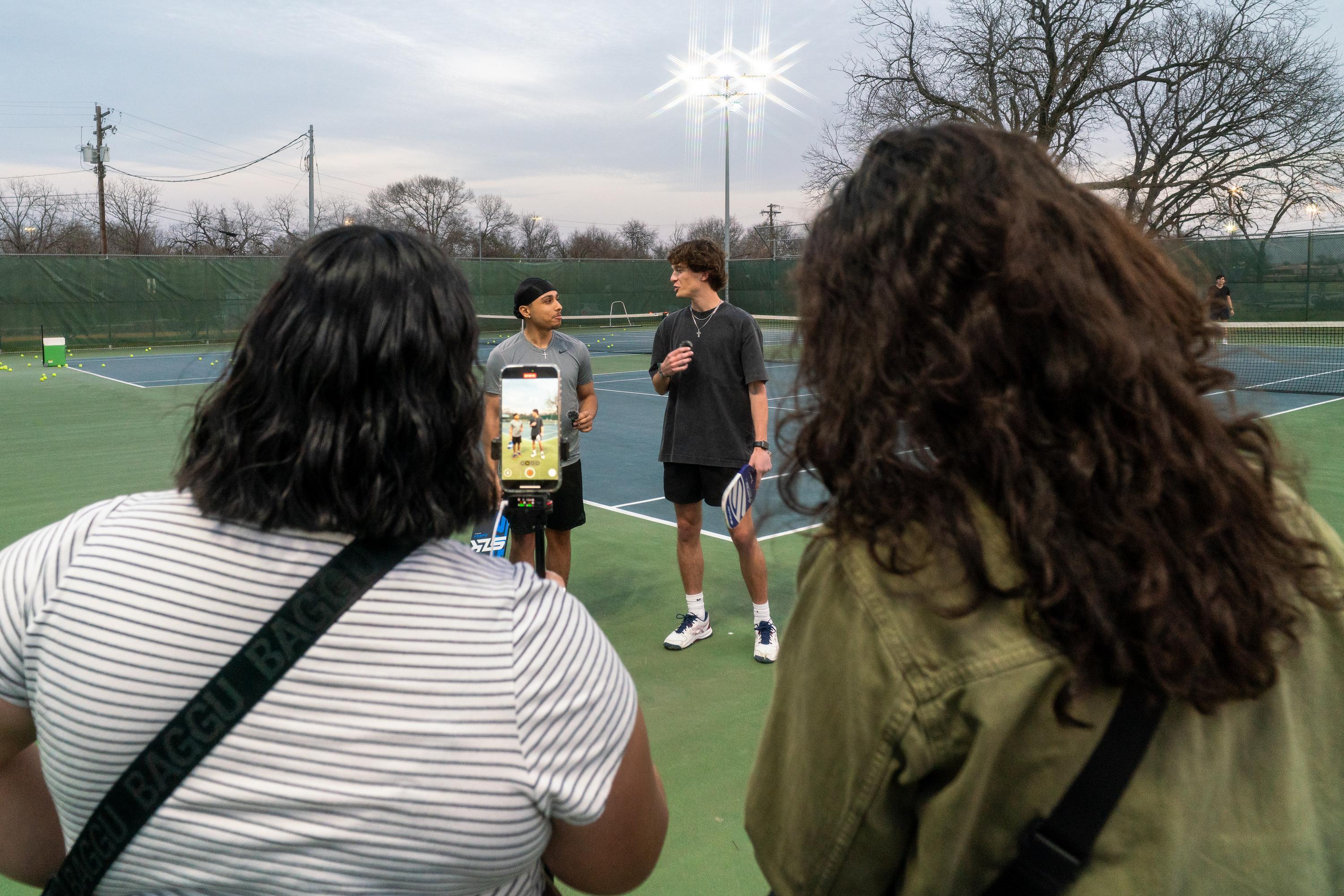 back of two women's heads as they film two men talking on a pickleball court
