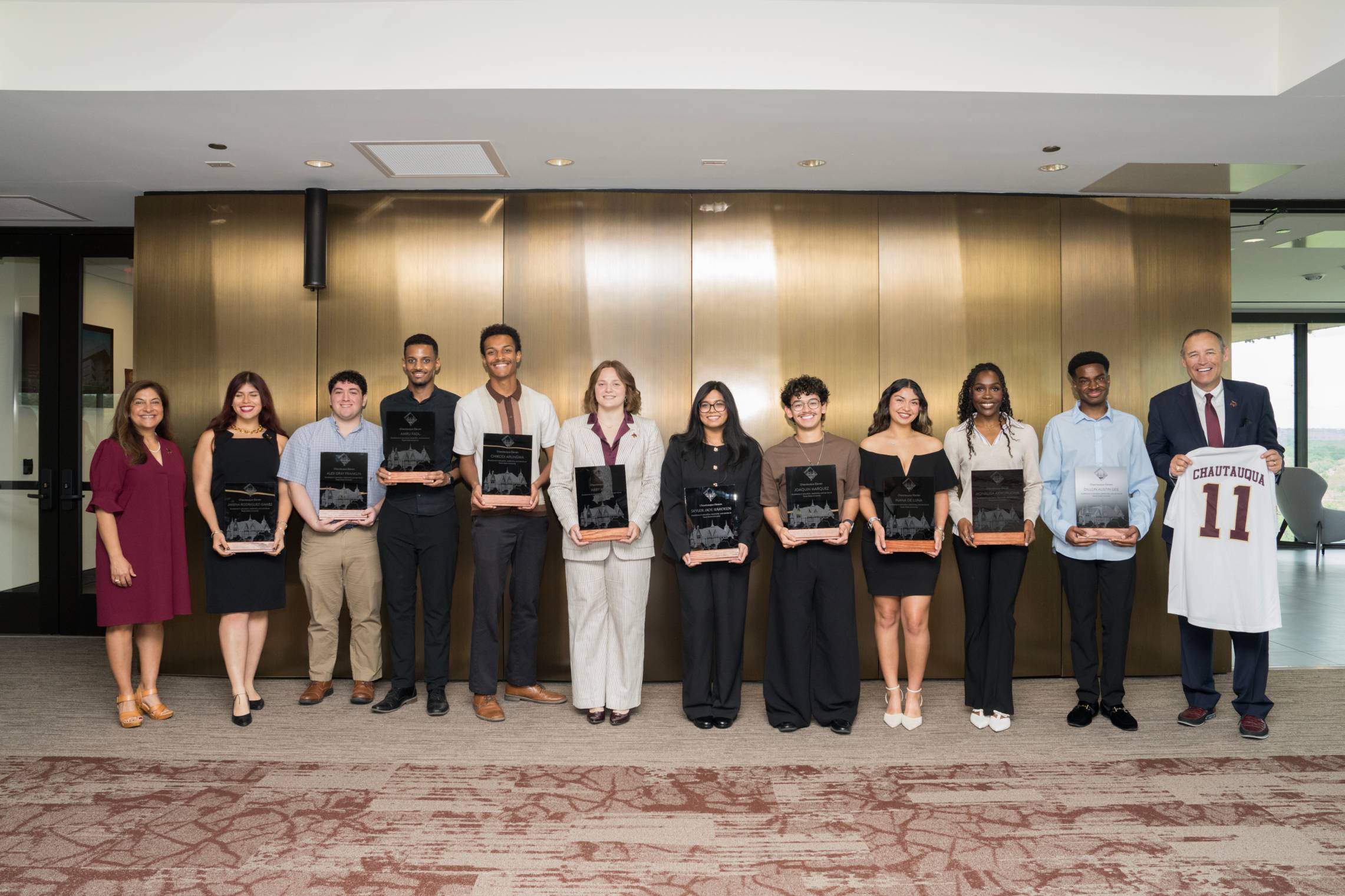 A group of eleven young adults and two older adults stand in a line indoors against a wood-paneled wall, each of the younger individuals holding a black award plaque. They are dressed in business or semi-formal attire and smiling at the camera. On the far right, a man in a suit holds up a white jersey with the number 11.