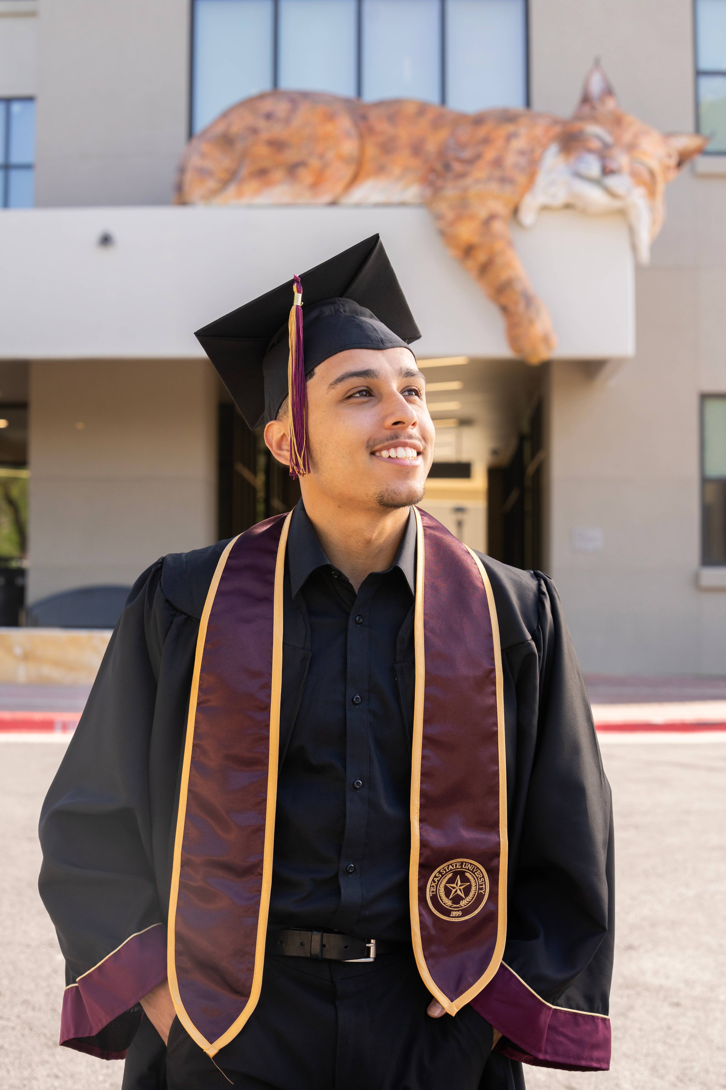 man in graduation regalia poses for photo in front of building with large sleeping bobcat sculpture on it