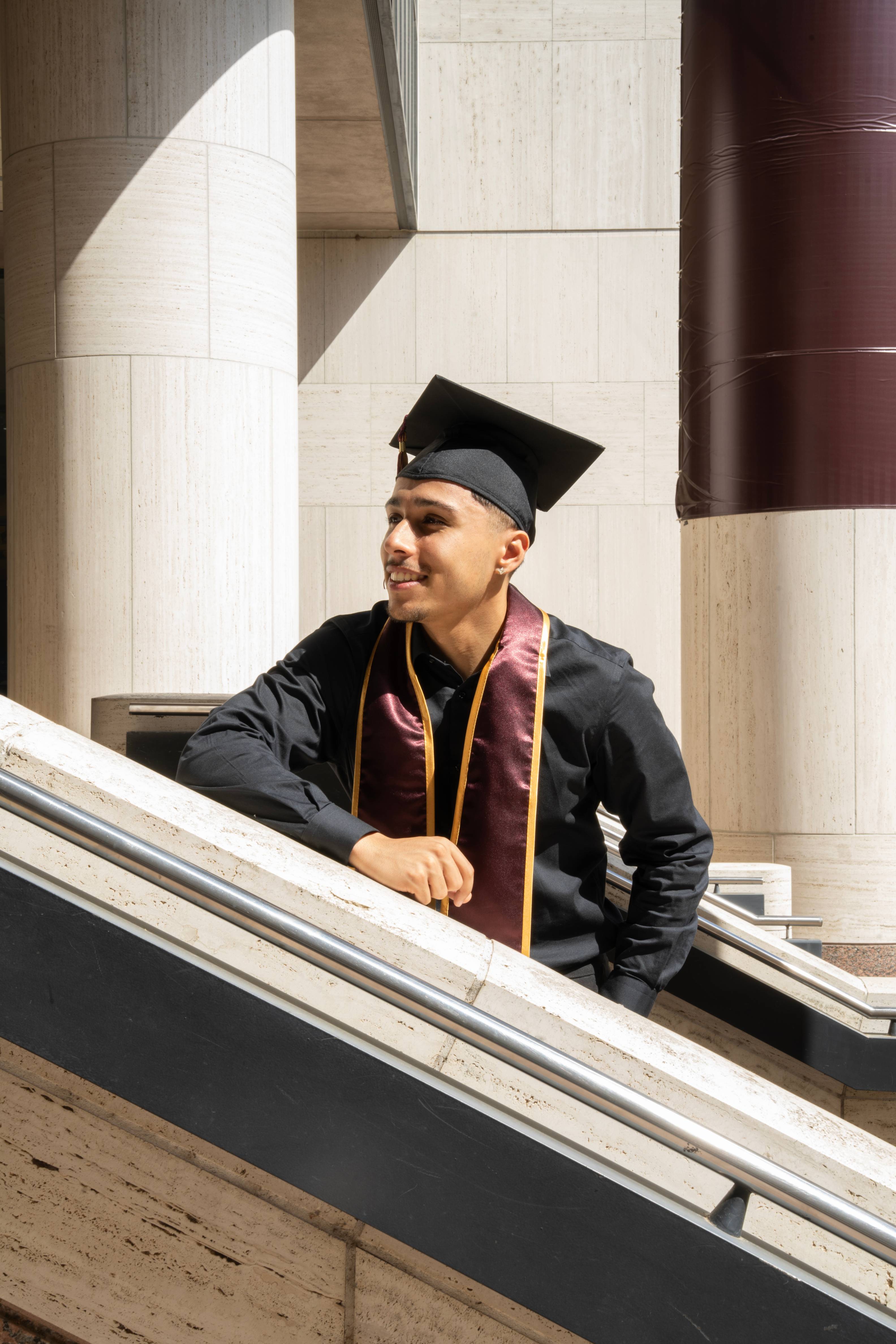 Graduate in cap and gown leans against the railing on the Alkek Library steps, looking out to the side with limestone columns behind him