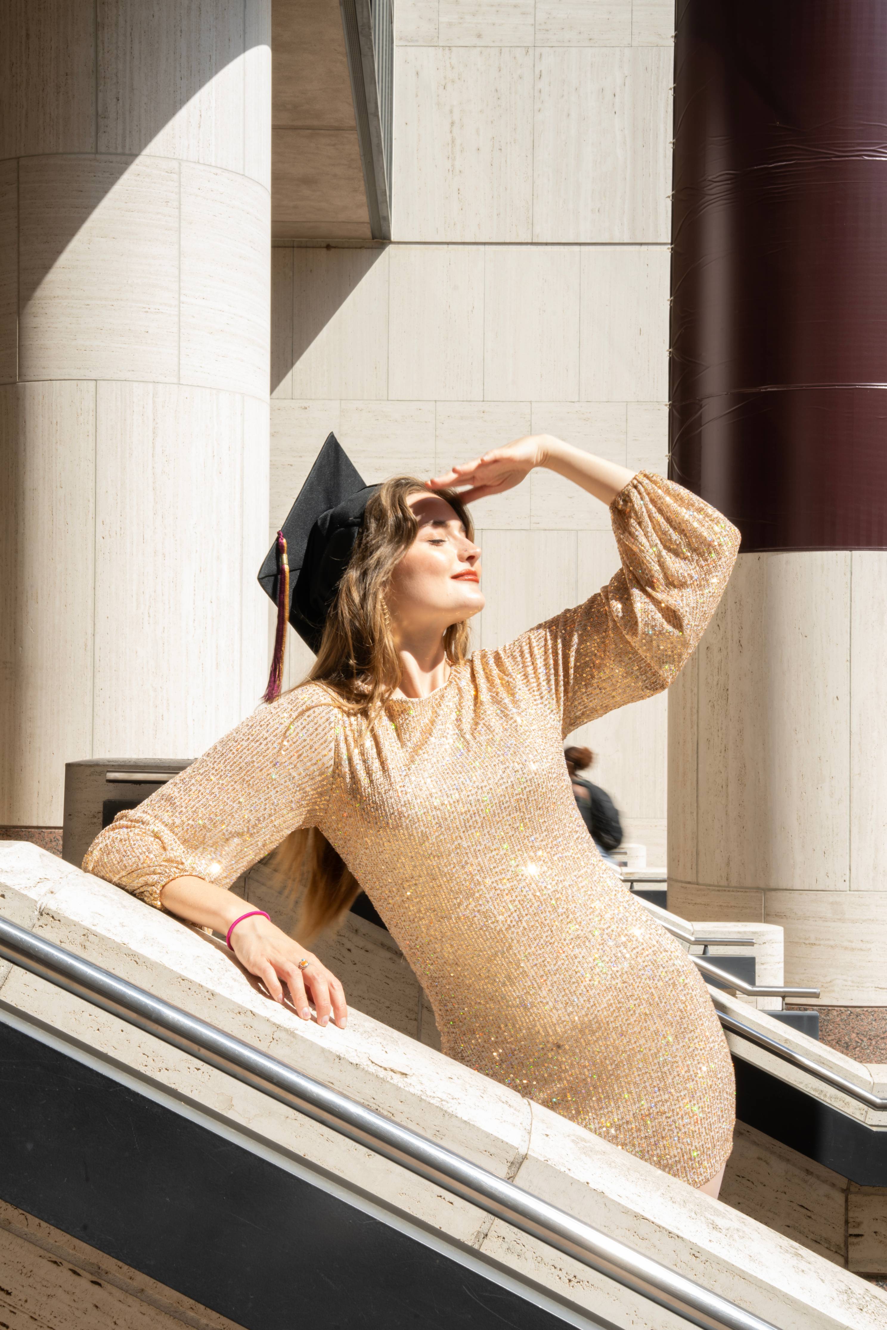 Graduate in a gold sequin dress leans on the Alkek Library railing, shielding her eyes from the sun and looking upward.