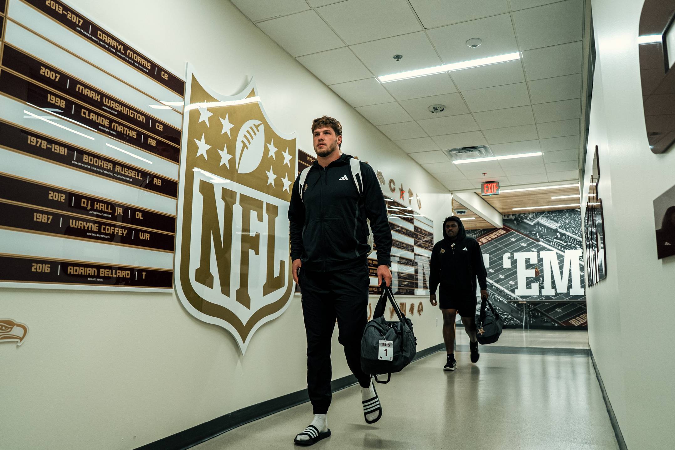 football players walk down a hallway adorned with names of former txst football players that have played in the NFL