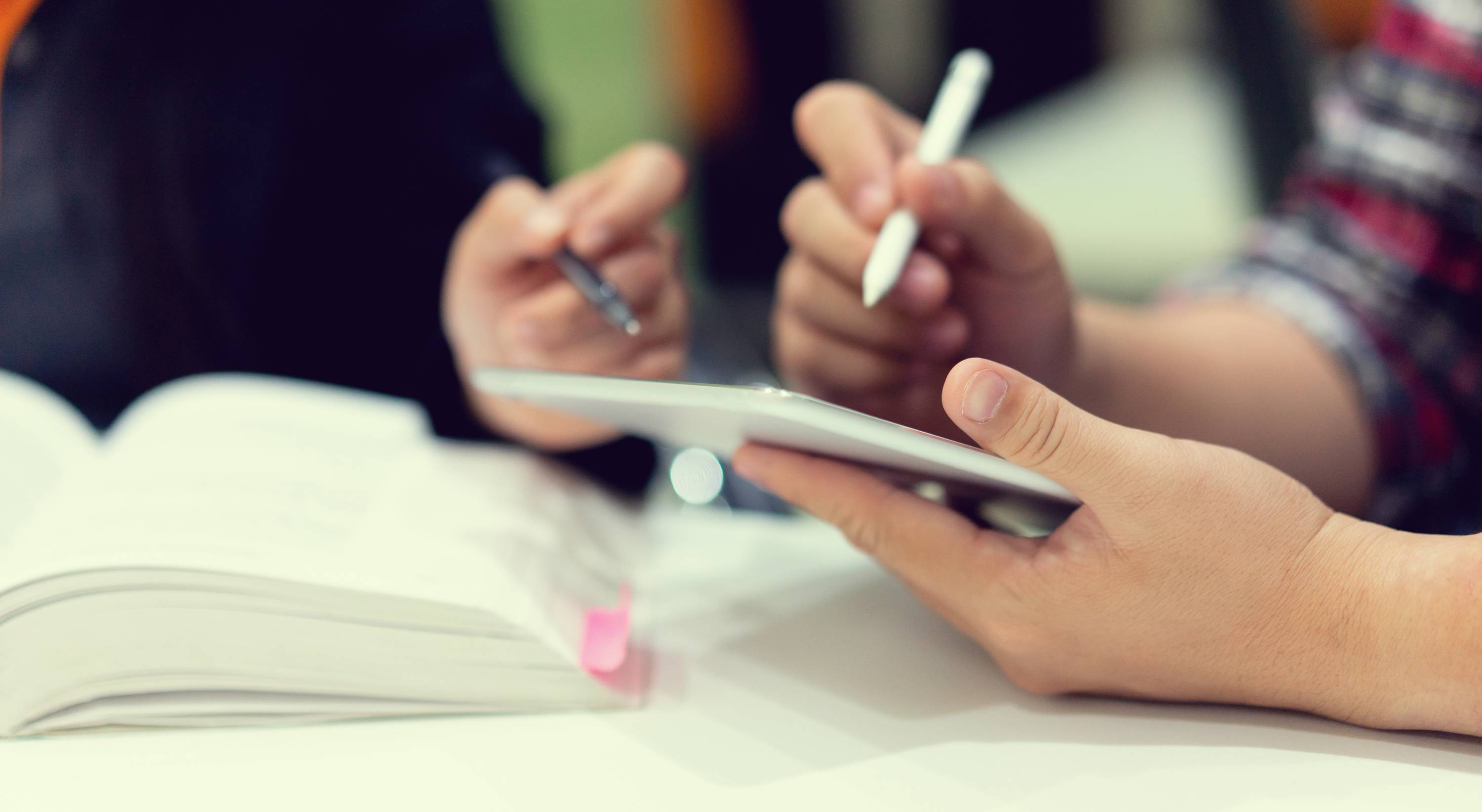 A close-up shot of two people collaborating at a desk. One person holds a tablet and a white stylus, while the person next to them points toward the screen with a pen. An open textbook with a pink bookmark sits in the foreground against a blurred background.