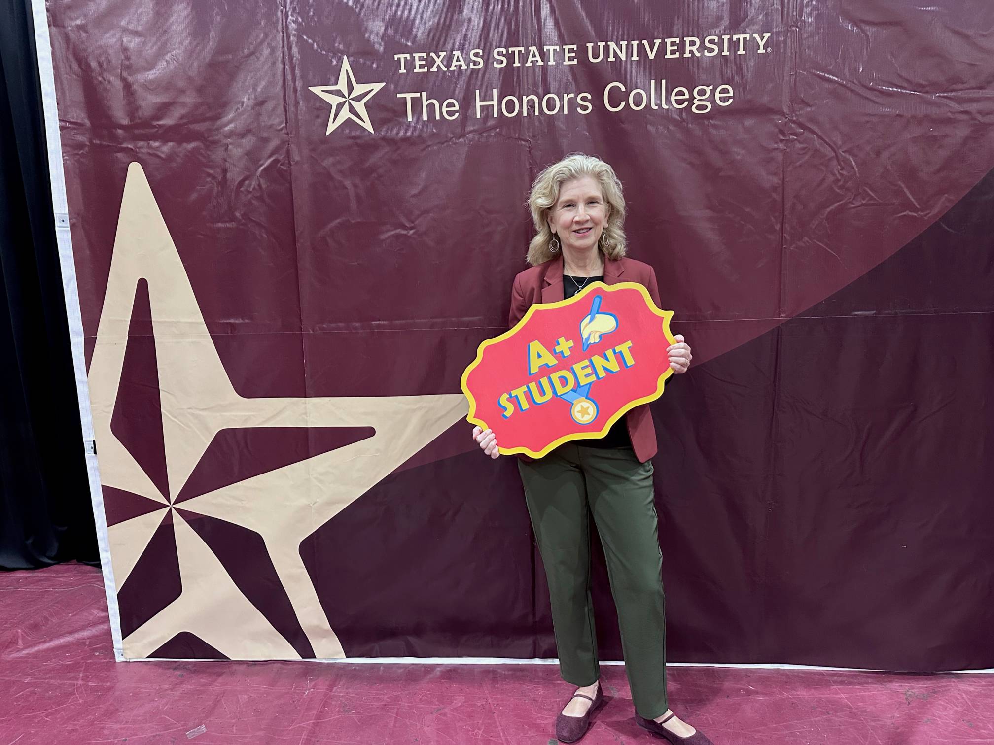 dean galloway holds up sign that reads "A+ Student" in front of Texas State University The Honors College sign
