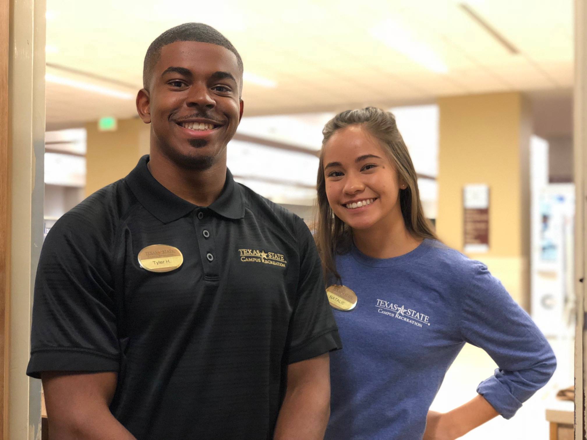 Two student employees wearing Texas State Campus Recreation shirts stand inside a campus facility, representing student employment opportunities.