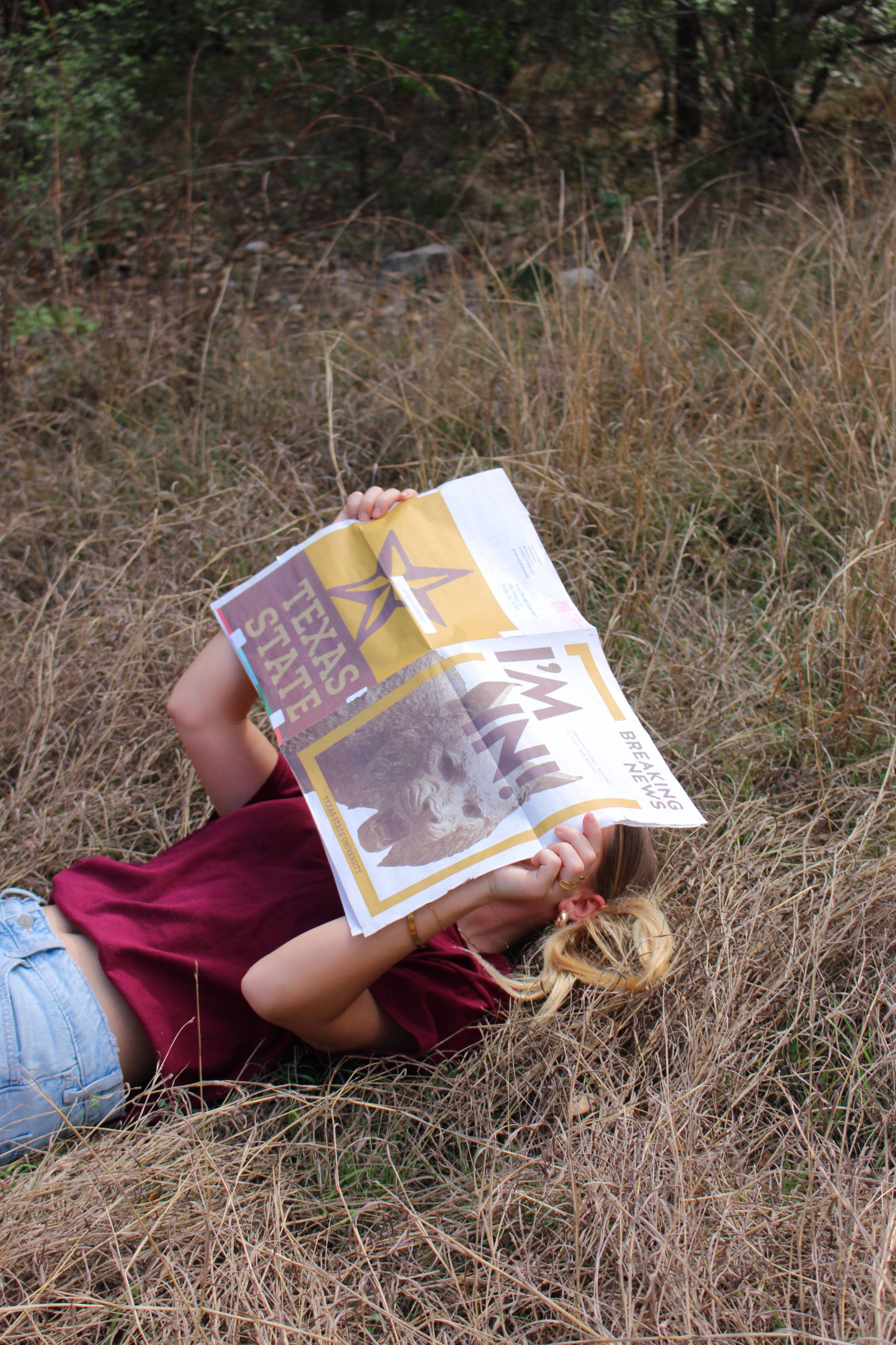 woman laying in grassy area while covering face with newspaper that reads "I'm in!"