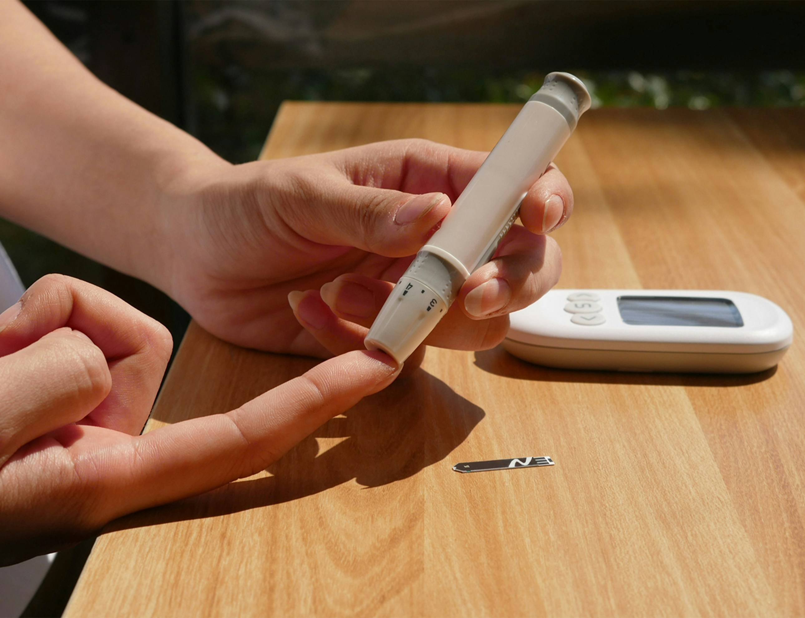 Close-up of a person using a lancing device to prick their fingertip for a blood glucose test, with a digital glucose meter and test strip on a wooden table nearby.